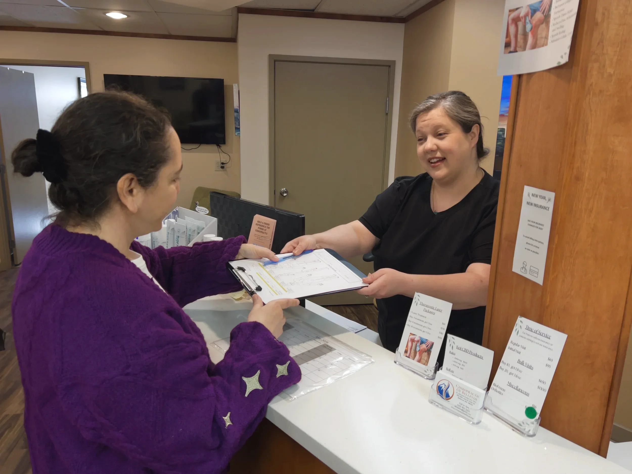 A woman with gray hair in a black shirt is handing a pamphlet to a woman with brown hair in a purple sweater at a reception desk. The desk has informational pamphlets, and a sign with health-related information is visible next to the woman. There is a black TV mounted on the beige wall behind them.
