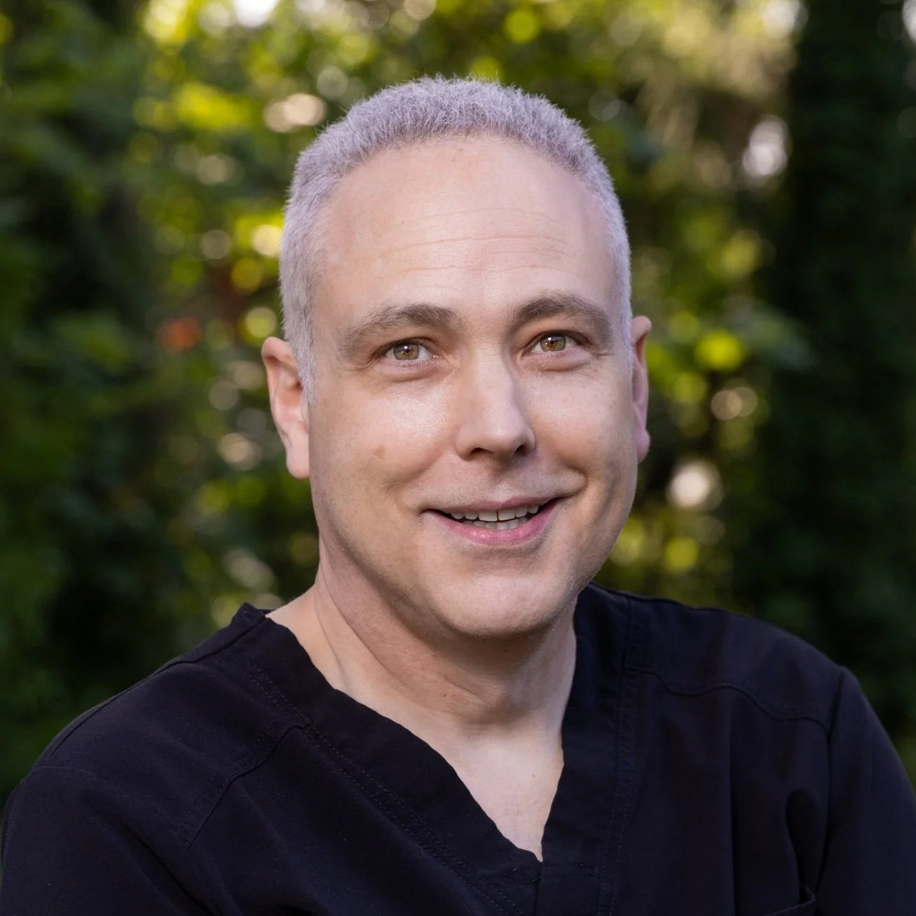 Portrait of a smiling man outdoors with blurred green foliage background.