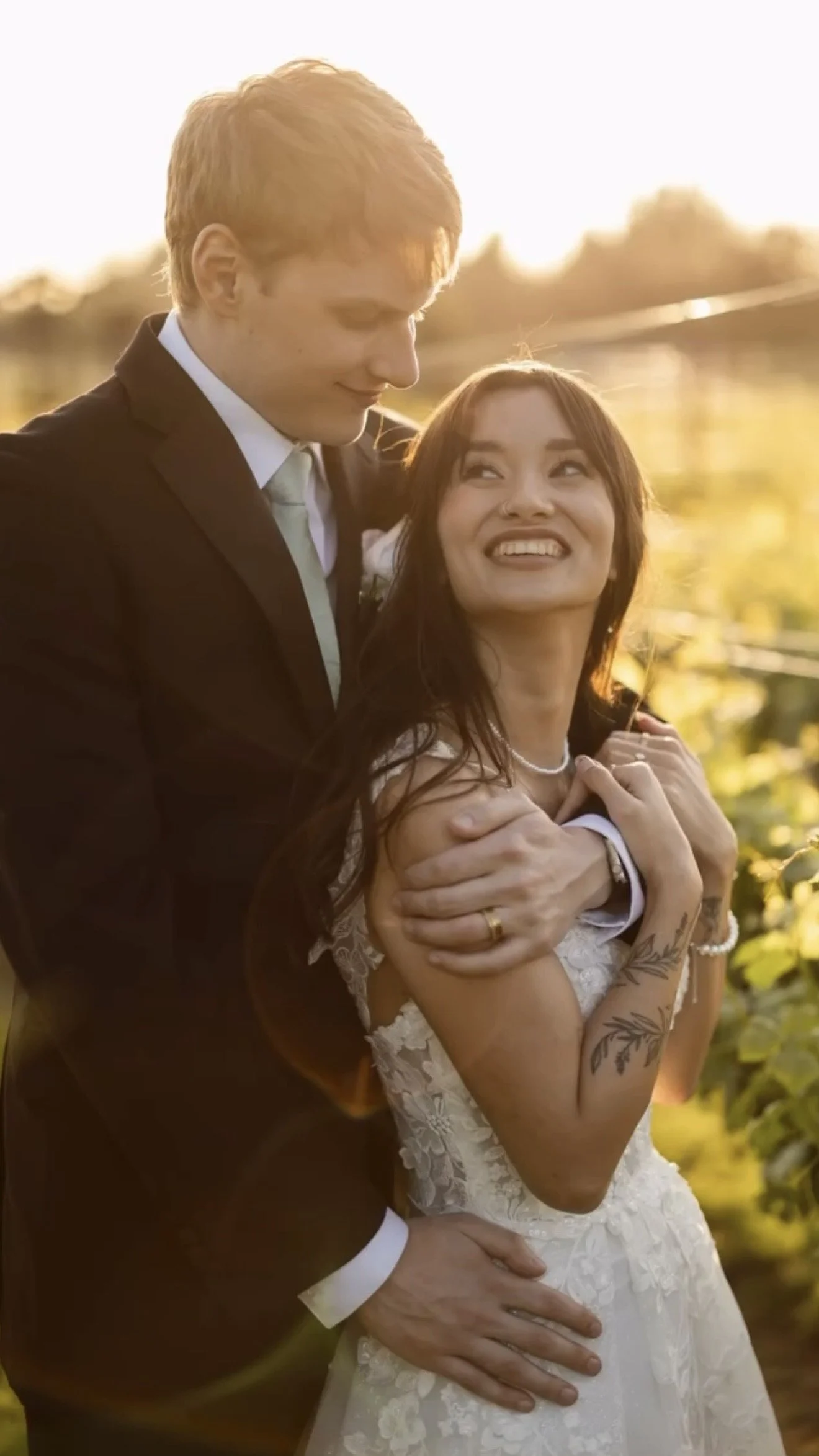 A happy couple celebrating their wedding outdoors during sunset, with the man in a black suit and the woman in a white lace wedding dress, smiling and embracing.