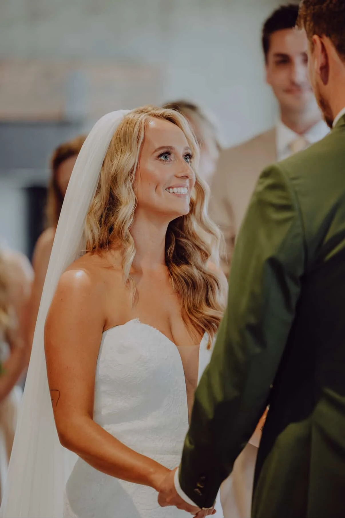 A bride with long, wavy blonde hair in a strapless white wedding dress, smiling at her groom during the ceremony, holding hands, with wedding guests in the background.