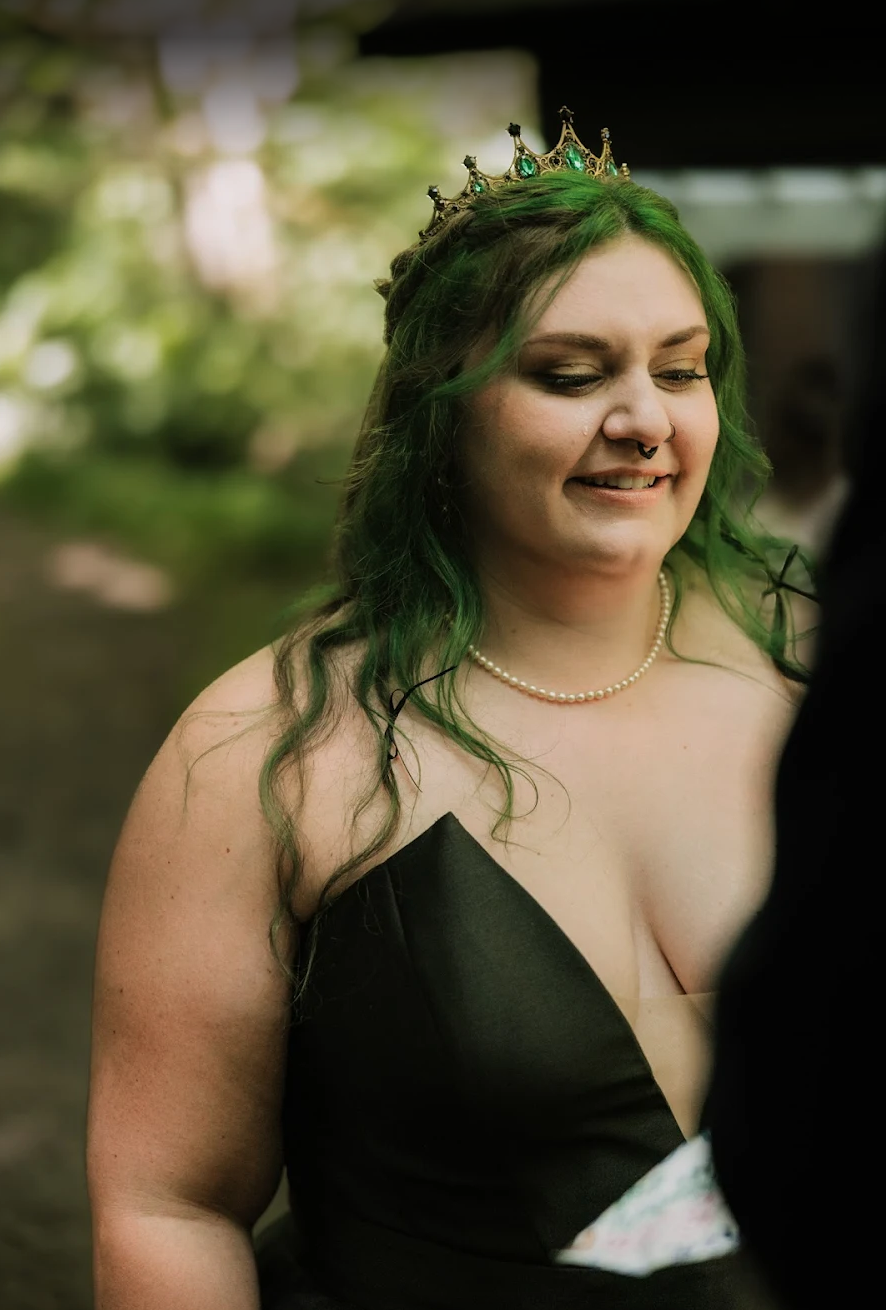 A woman with green hair wearing a tiara, pearl necklace, and black dress, smiling and looking downward at her wedding