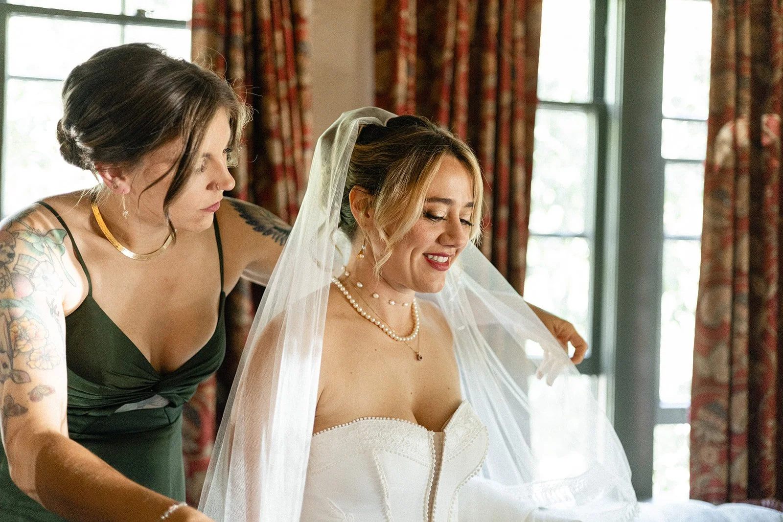 A bride in a white wedding dress and pearl necklace smiling as she is helped by another woman, possibly her stylist or friend, in a room with large windows and floral curtains.