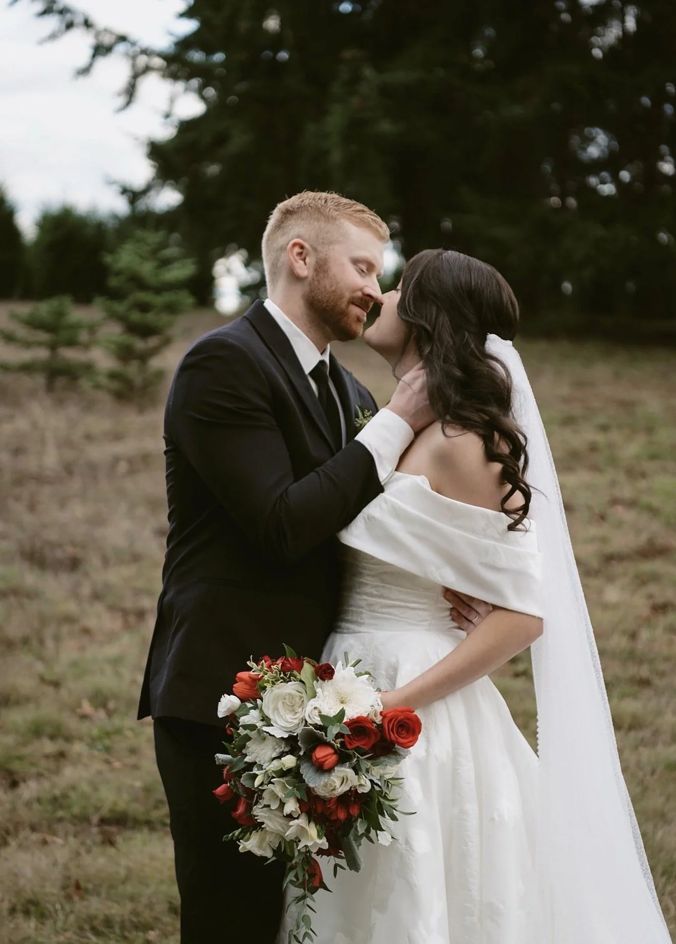 A newlywed couple sharing a romantic moment outdoors, with the man in a black suit and tie and the woman in an off-shoulder white wedding dress holding a bouquet of red and white flowers.