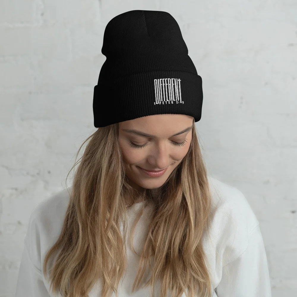 A woman with long blonde hair wearing a black knit beanie with a white barcode on it, looking down with a smile, against a white brick wall background.