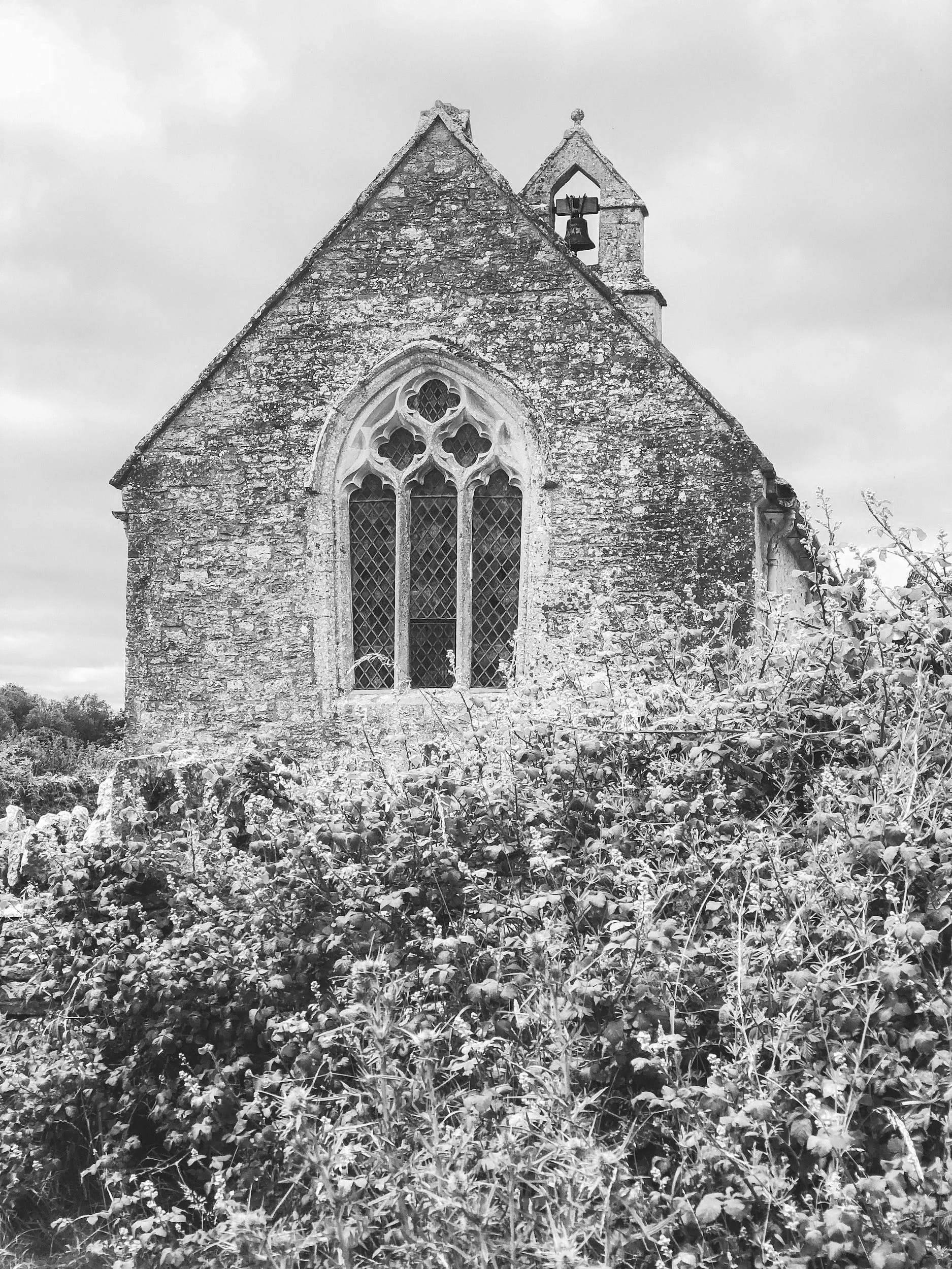 An old stone church with a large stained-glass window and a small bell tower, surrounded by overgrown bushes and plants, under a cloudy sky.