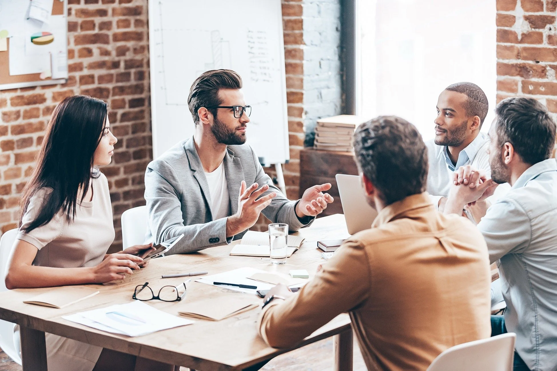 Business meeting with five individuals seated around a wooden table in a brick-walled office, engaged in discussion. One man in a gray blazer and glasses is speaking, while others listen attentively. The table has documents, notebooks, a pair of glasses, pens, and a glass of water.