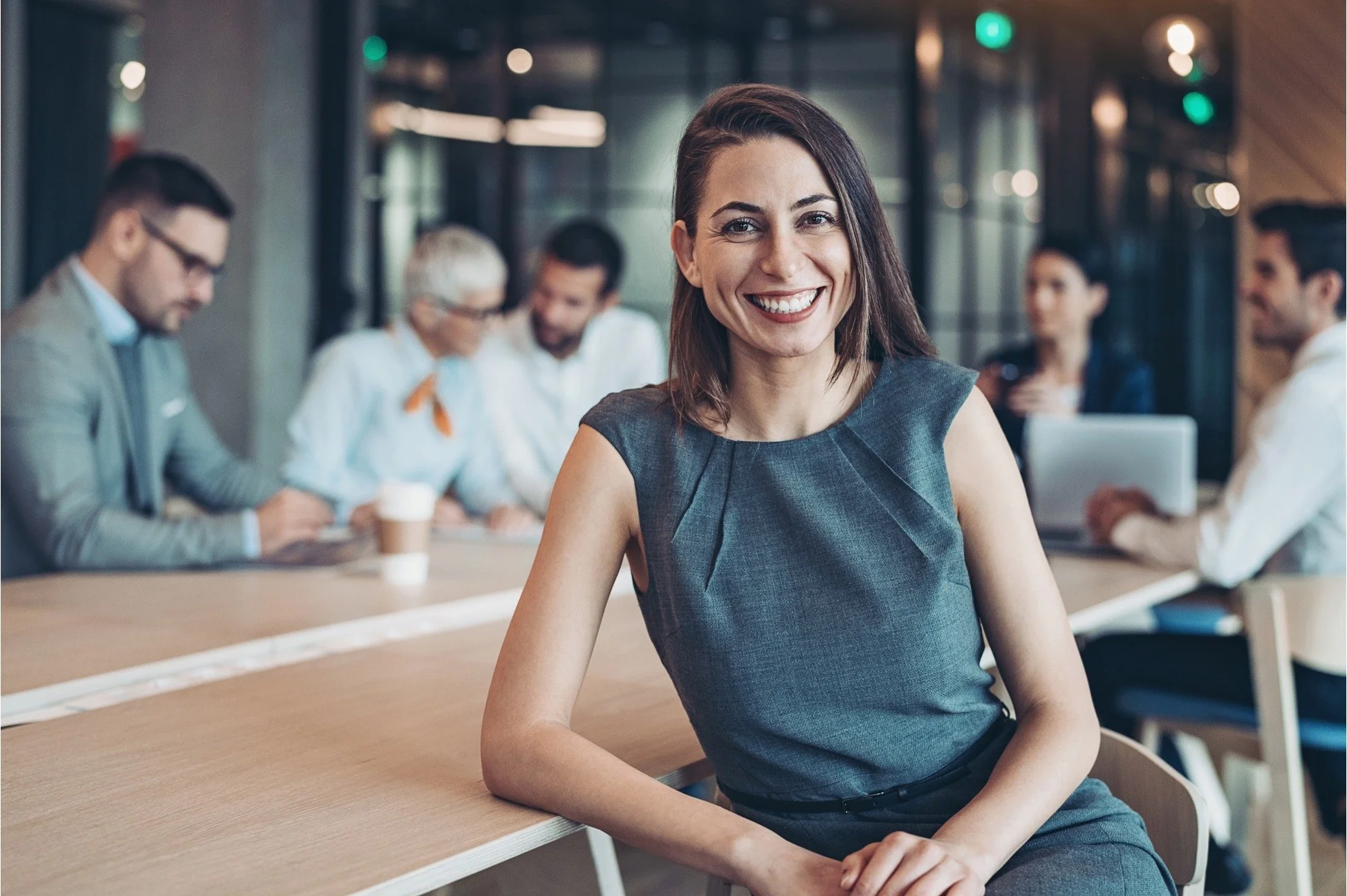 Woman smiling at the camera sitting at a table in a modern office conference room with colleagues working in the background.