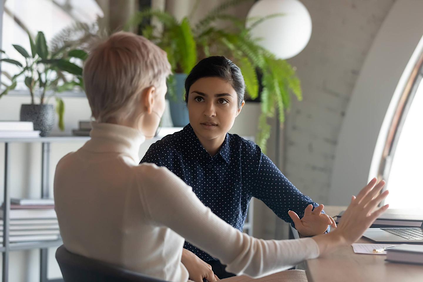 Two women having a serious conversation in an office with green plants in the background.