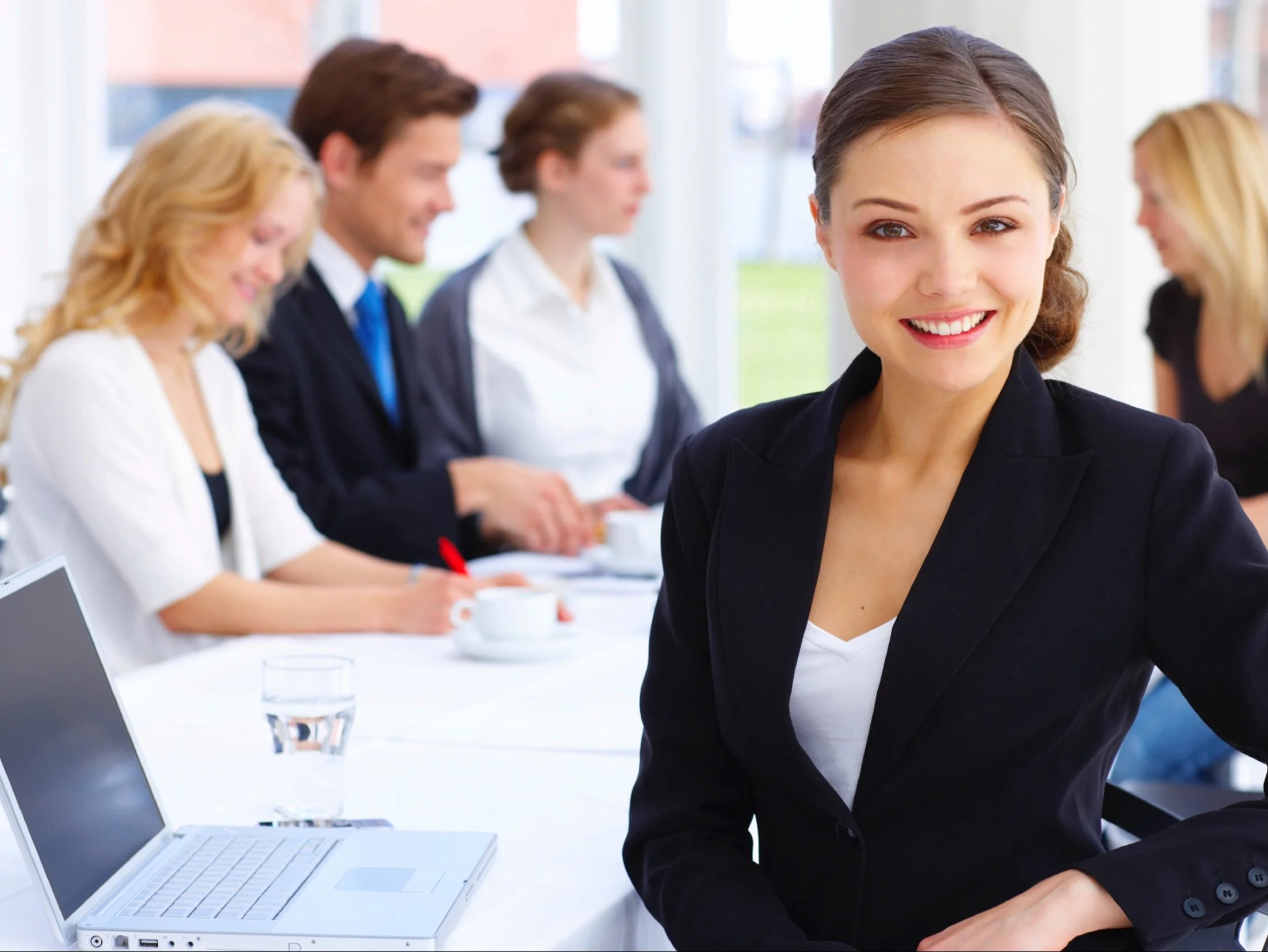 Business meeting with five diverse adults seated around a table, woman in black blazer smiling at the camera, laptop, glass of water, and cups on the table.