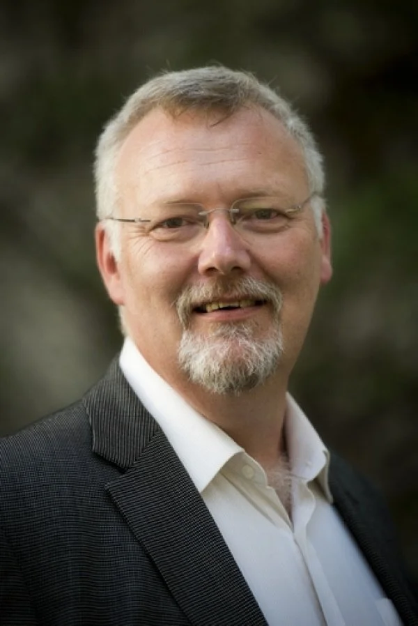 A middle-aged man with glasses, a beard, and mustache, wearing a white collared shirt and a dark blazer, smiling outdoors with a blurred natural background.