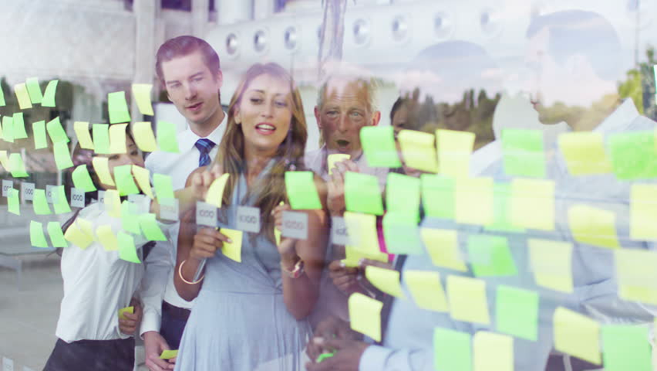 Group of businesspeople gathered around a glass wall covered with colorful sticky notes, engaged in a collaborative planning or brainstorming session.