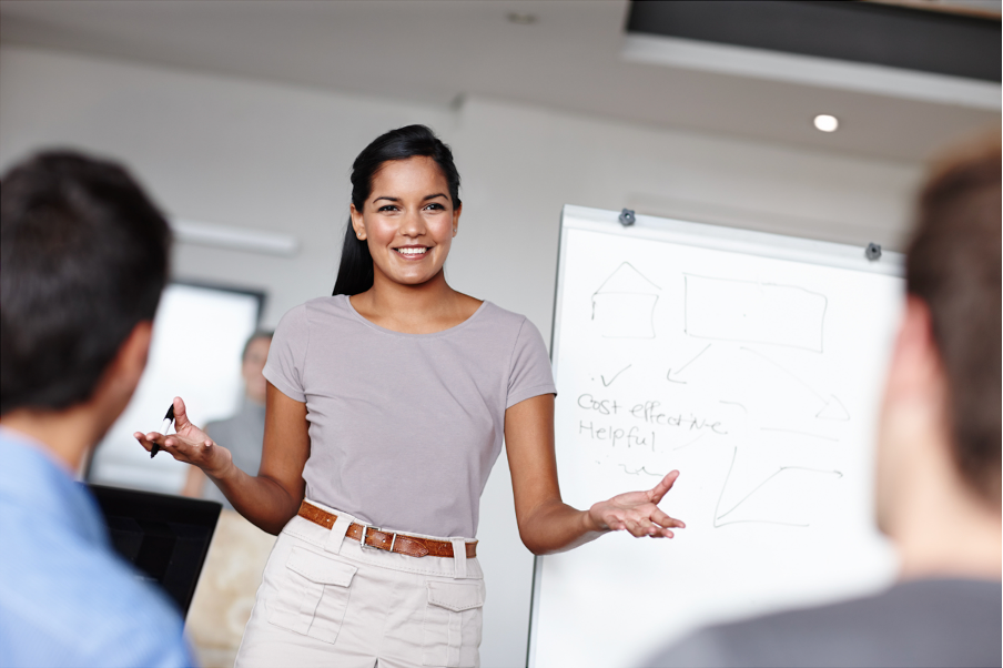 A woman in a purple t-shirt and beige pants is giving a presentation in front of a whiteboard, smiling while speaking to a group of people in an indoor setting.