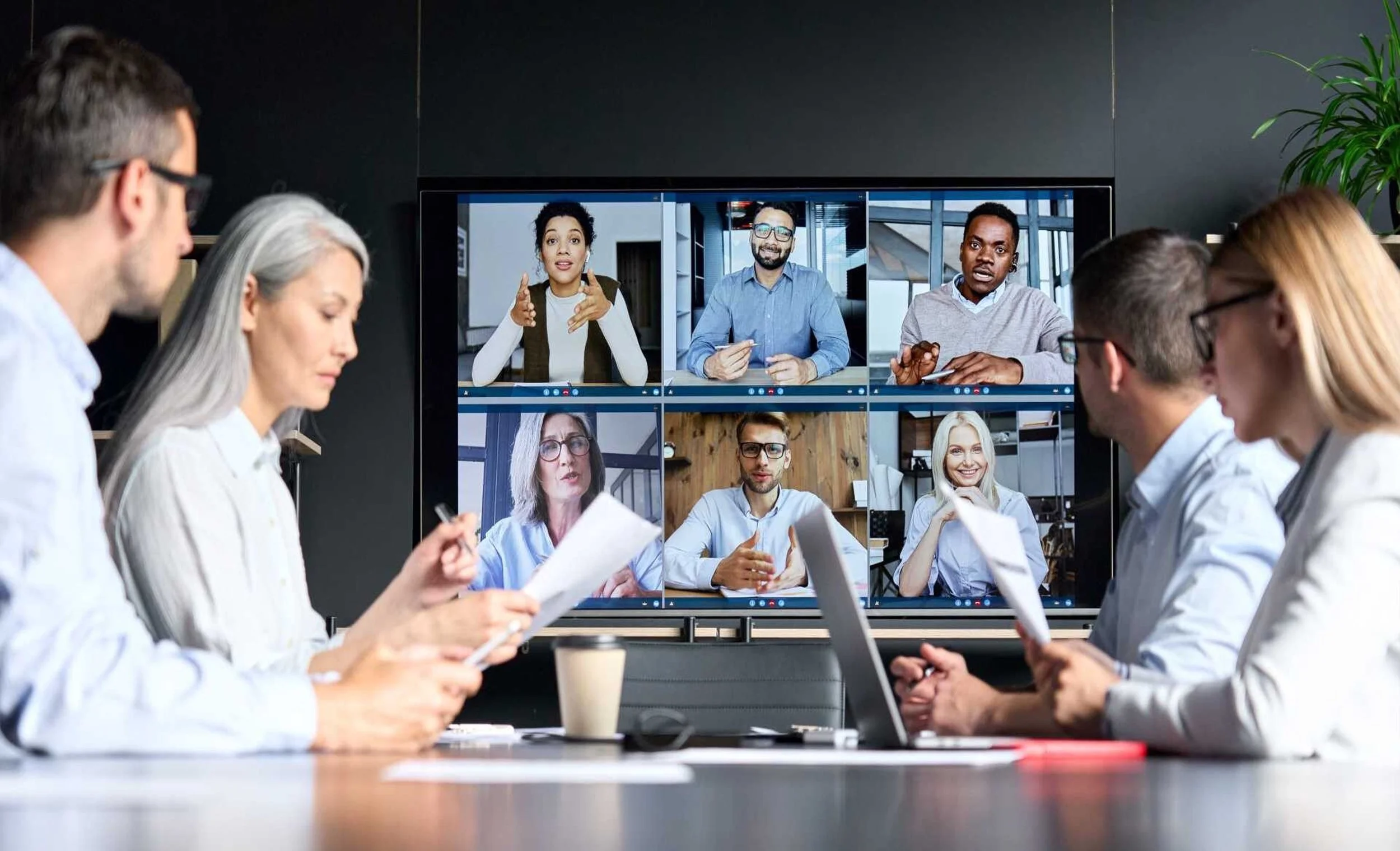 People in a conference room participating in a virtual meeting, with nine video call participants displayed on a large screen.