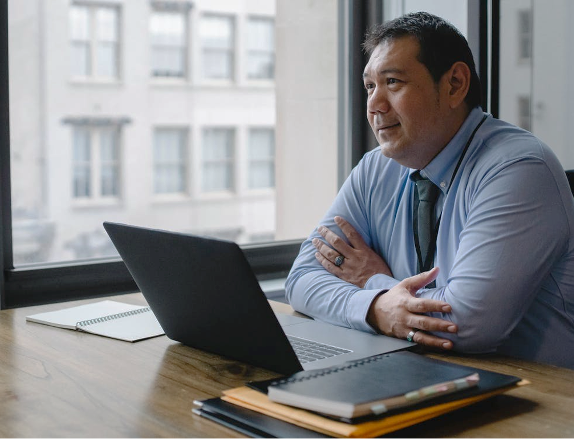A man in a blue shirt and tie seated at a desk with a laptop, notebooks, and a notepad, looking out the window.