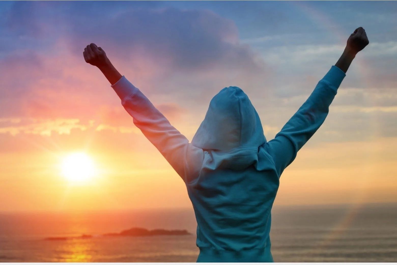 Person wearing a hoodie with arms raised during sunset at the beach.