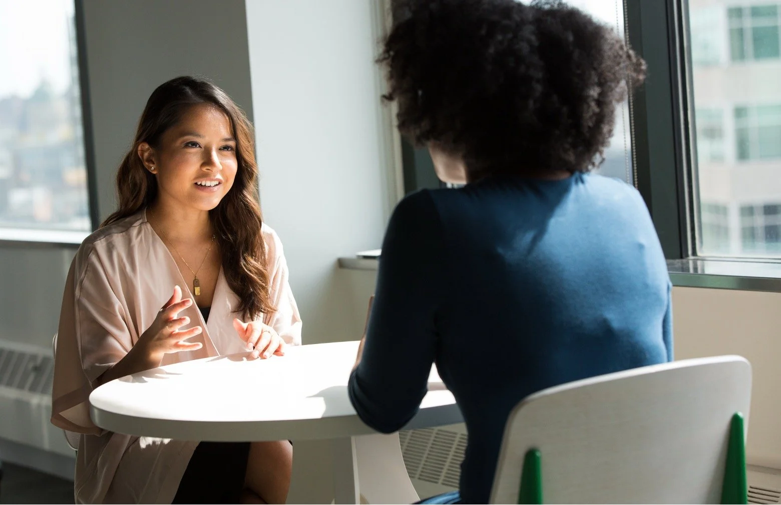 Two women seated at a white table having a conversation in an office with large windows.