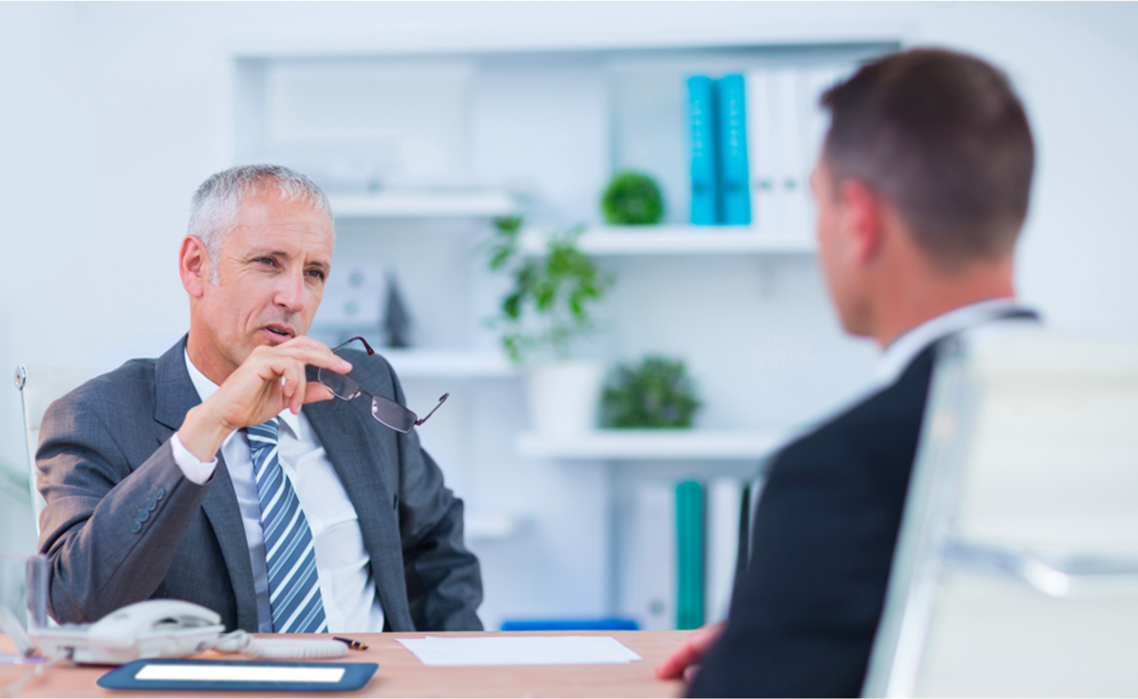 Older man in suit holding glasses and talking to younger man in an office setting.