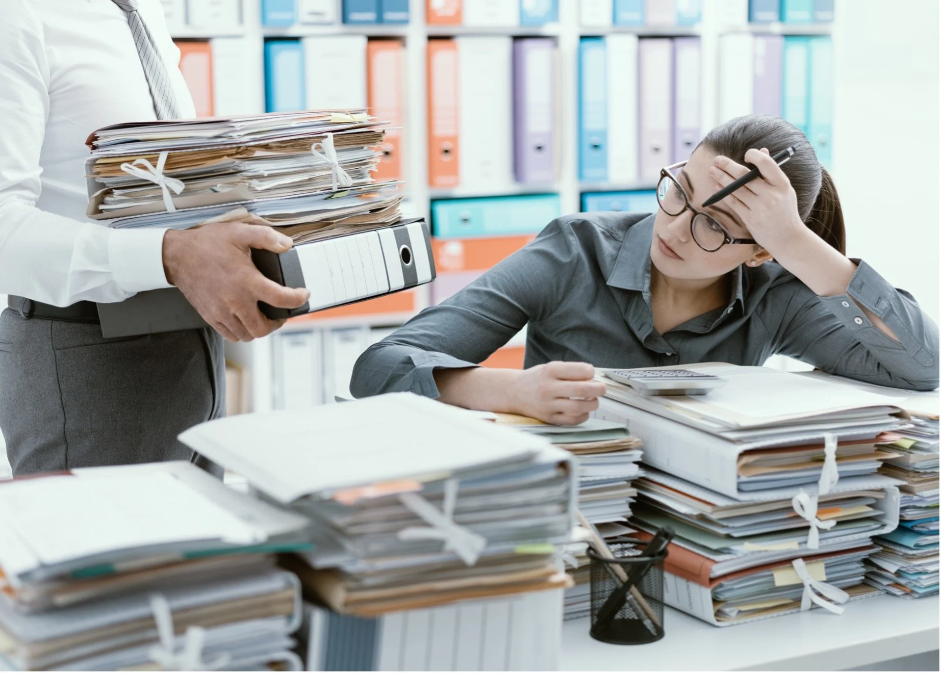 A woman working at a cluttered desk with stacks of files and documents, looking stressed, while a man handing her a thick folder in an office environment.