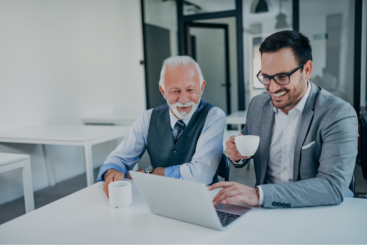 Two men in business attire smiling and looking at a laptop in a modern office. The older man has white hair and a beard, and the younger man has dark hair and glasses. Both are holding coffee mugs.