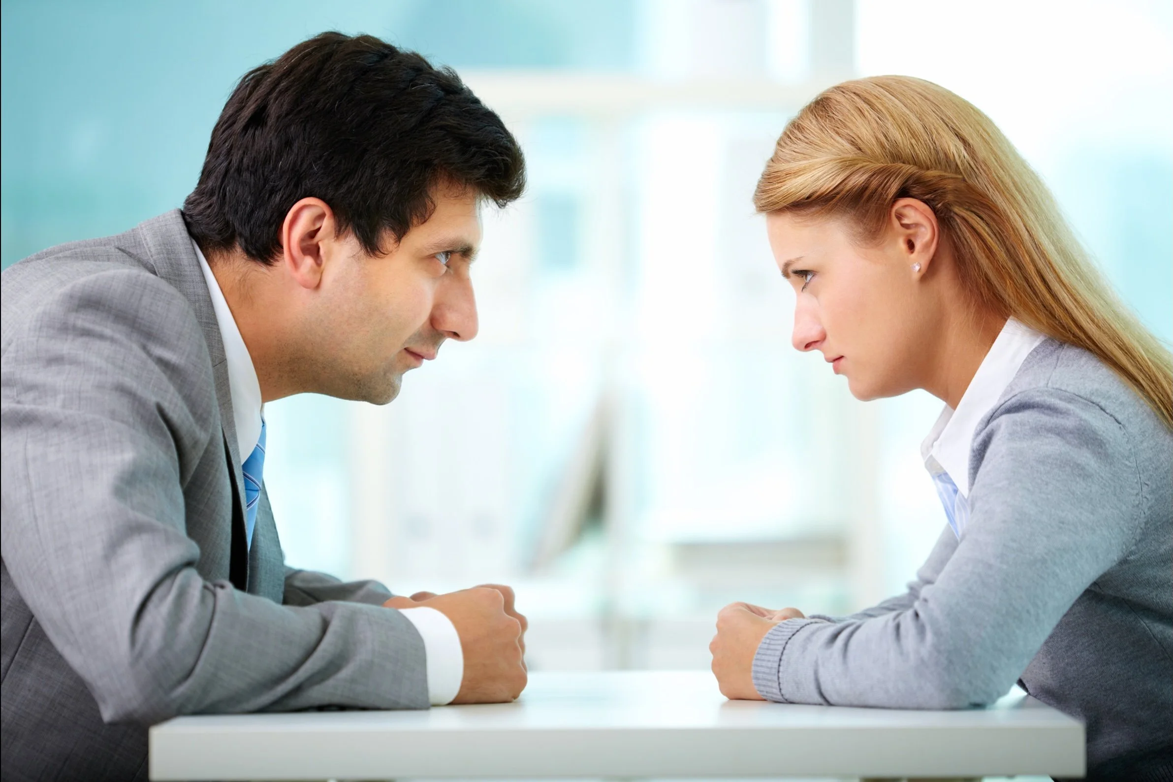 A man and woman sitting at a table facing each other with intense expressions, leaning forward with their foreheads almost touching, in a confrontational or serious discussion.