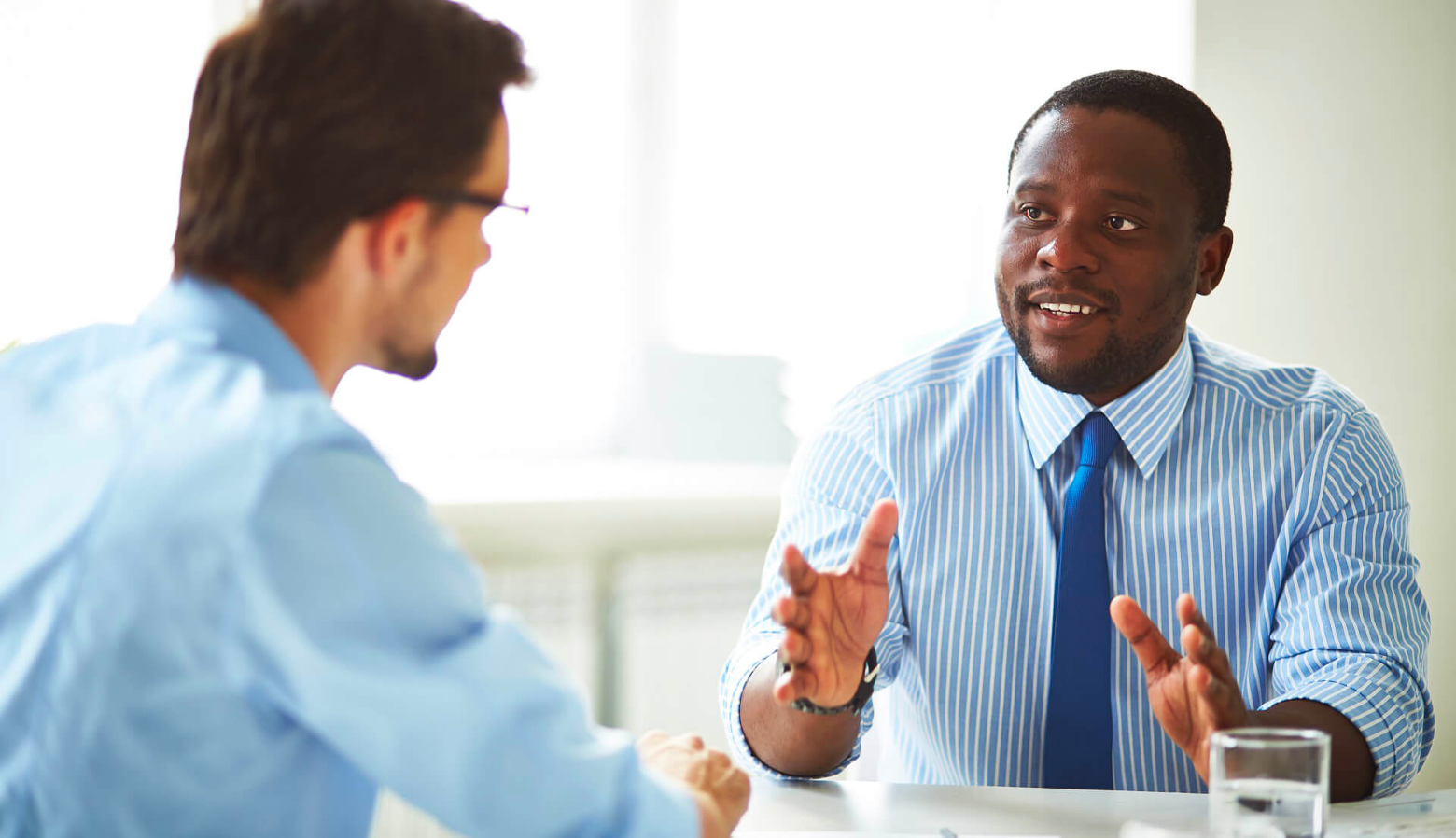 Two men having a conversation at a table, one is speaking and gesturing with hands, the other is listening. Both are dressed in business attire.