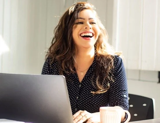 A woman with long brown hair, wearing a dark polka dot top, is sitting at a table, smiling and laughing while looking to the side. There is a laptop and a cup in front of her.