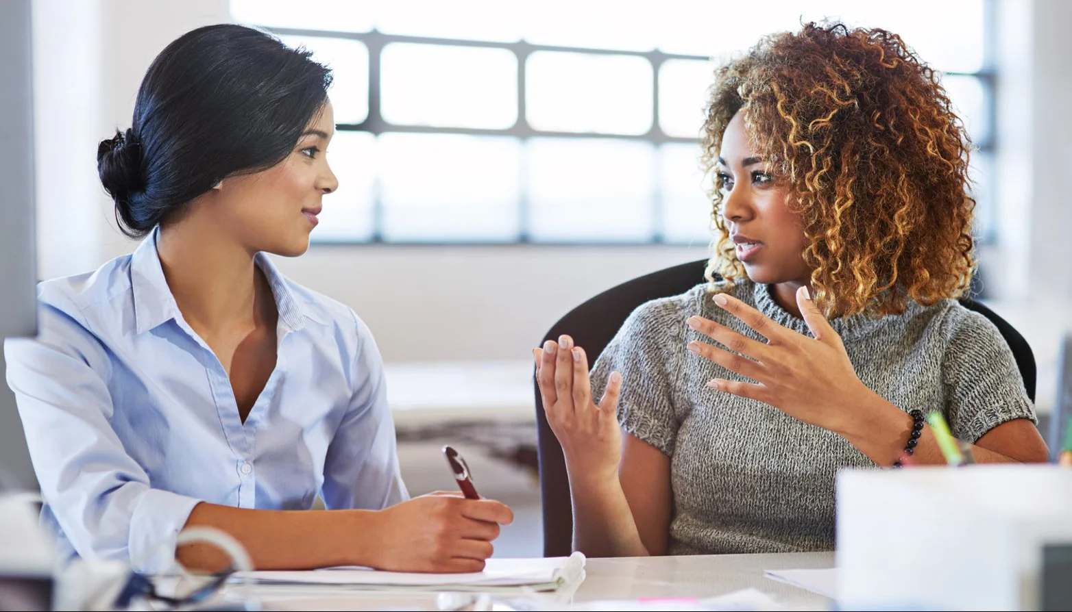 Two women sitting at a desk having a discussion, one explaining something, in a bright office space