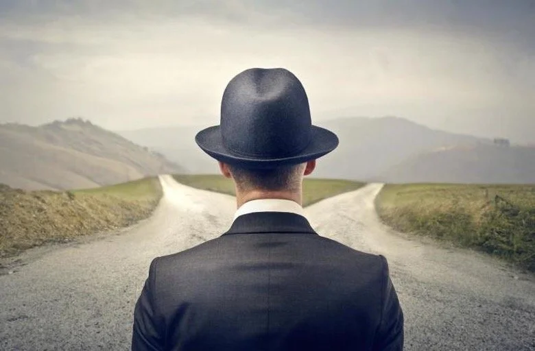 Back view of a man in a suit and hat looking at two diverging dirt roads in a rural landscape.