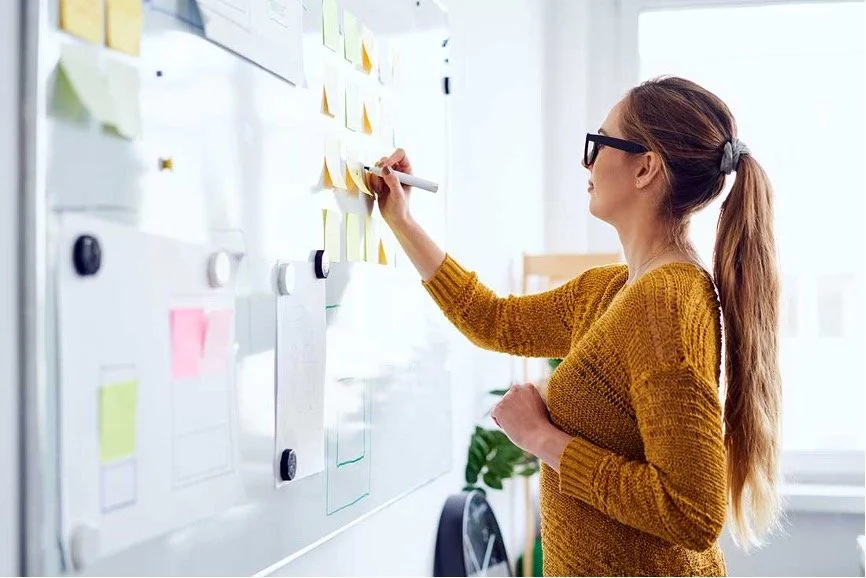 A woman with glasses and a ponytail writes notes on a whiteboard covered with colorful sticky notes in a bright, modern office.