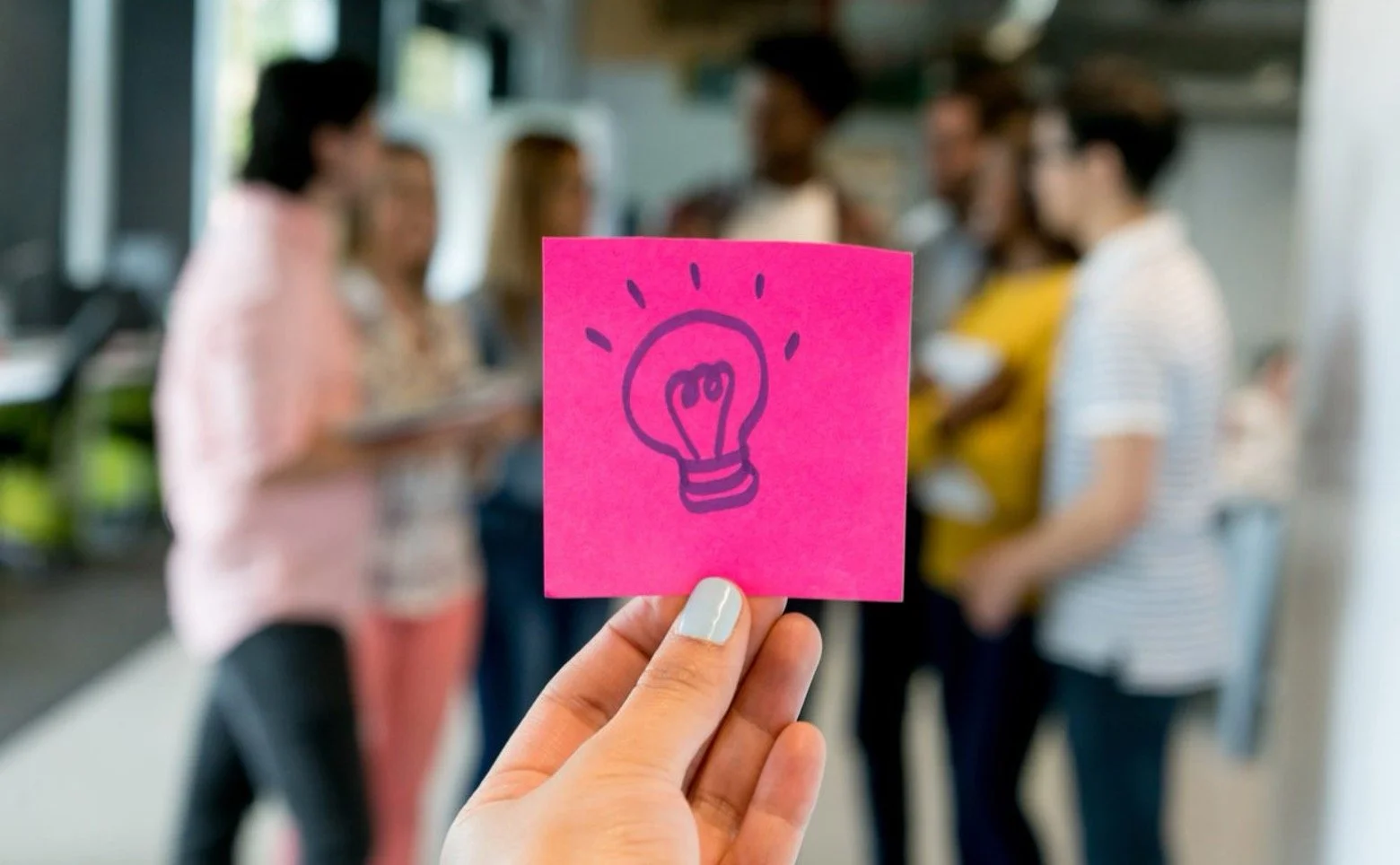 Person holding a pink sticky note with a hand-drawn lightbulb, with a group of blurred people in the background.