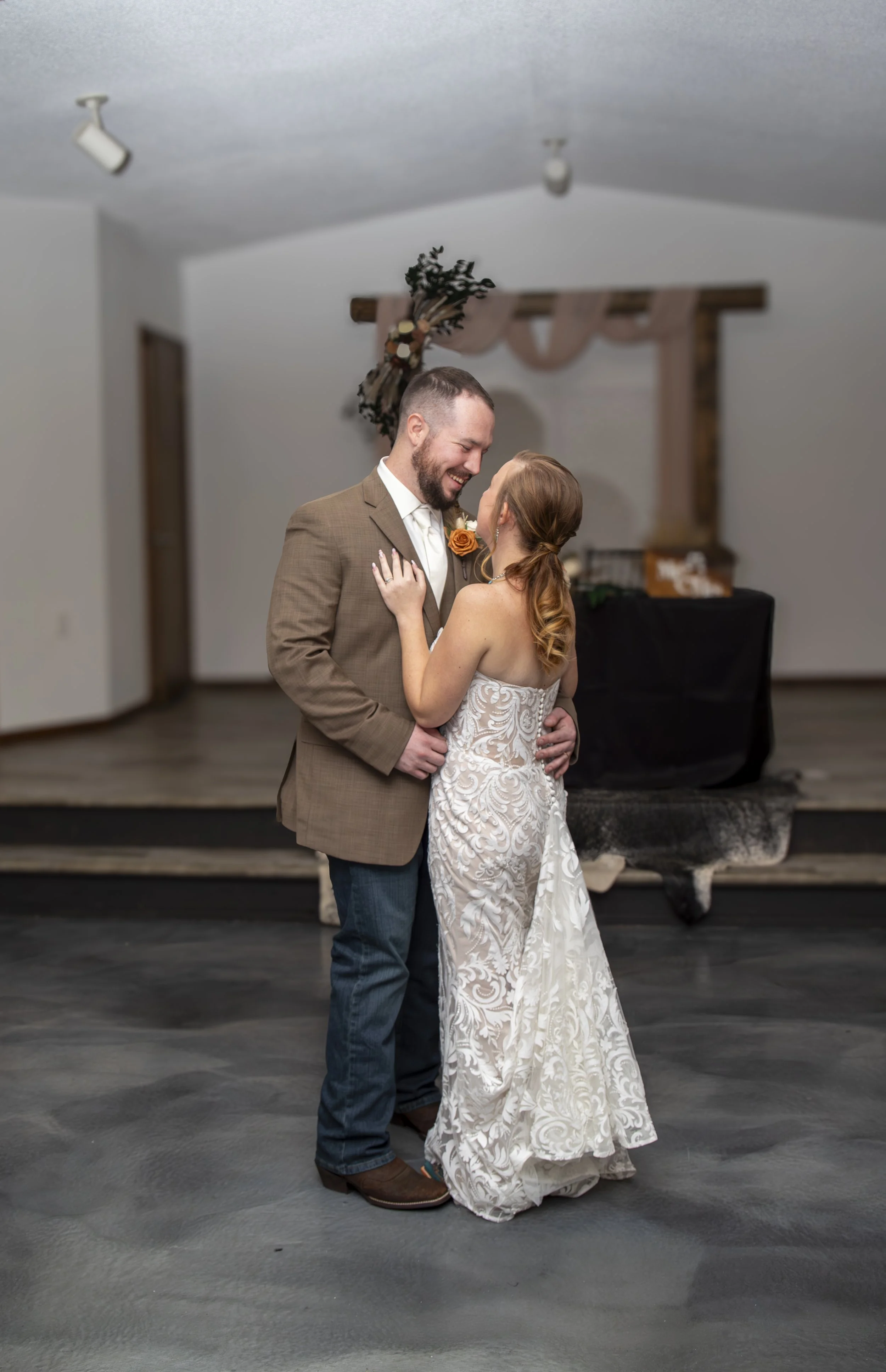 A couple embracing at a wedding, with the bride in a white lace dress and the groom in a brown jacket and jeans, standing indoors near a decorated arch.