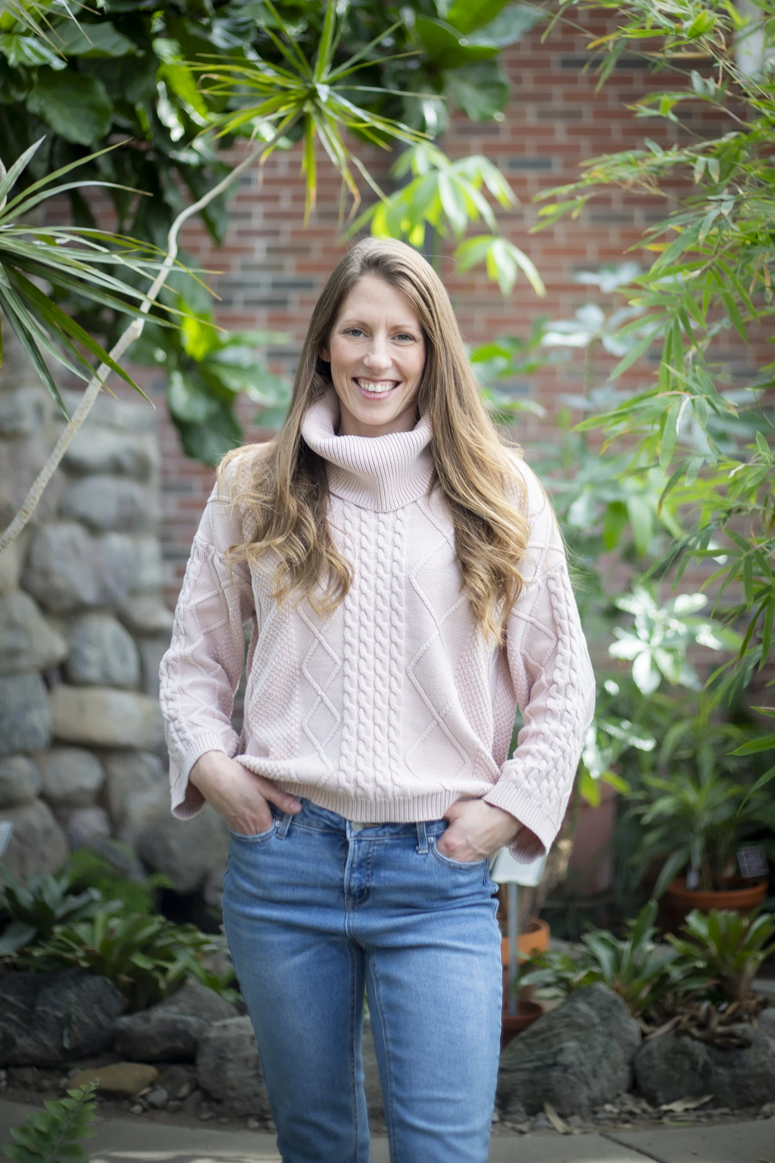 A person wearing a light pink sweater and blue jeans standing outdoors with green plants and a stone wall in the background.