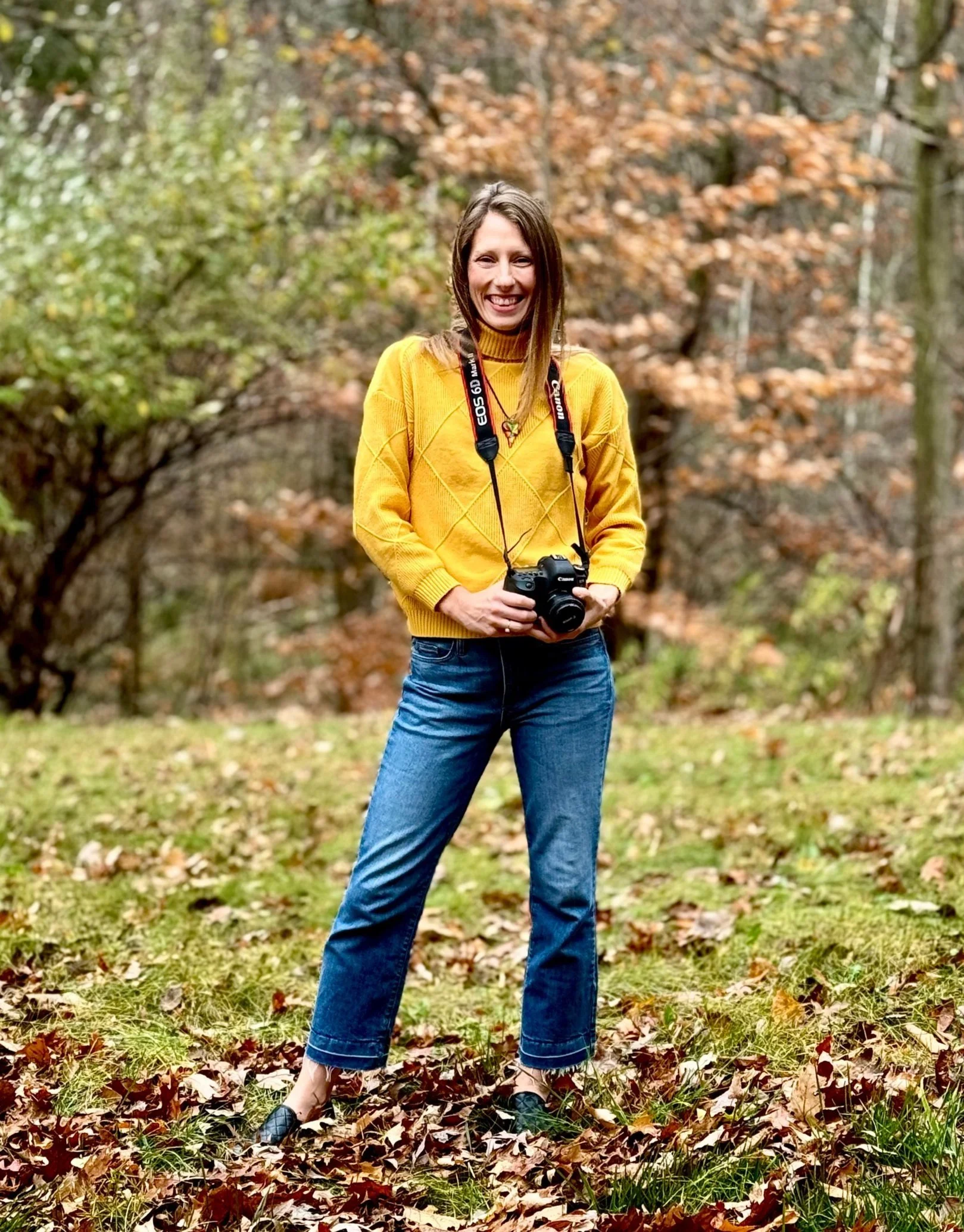 A person standing outdoors in a park, wearing a yellow sweater, jeans, and holding a camera. The surroundings are grassy with fallen leaves and trees in the background.