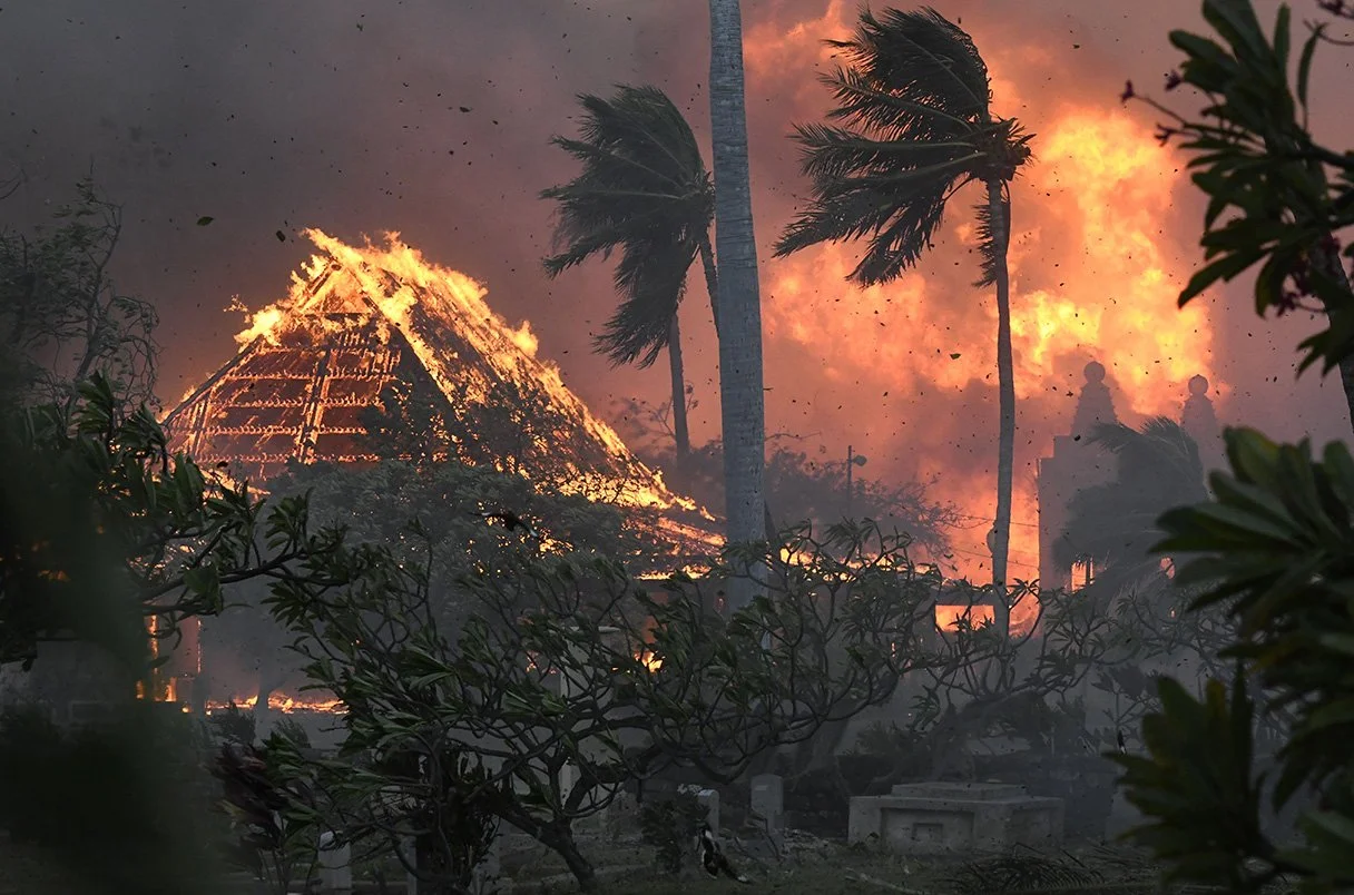 A house is on fire, with flames and smoke rising into the sky. Several palm trees are visible, and the scene appears to be in a tropical area. The sky is filled with orange and dark clouds, indicating a wildfire or large blaze.