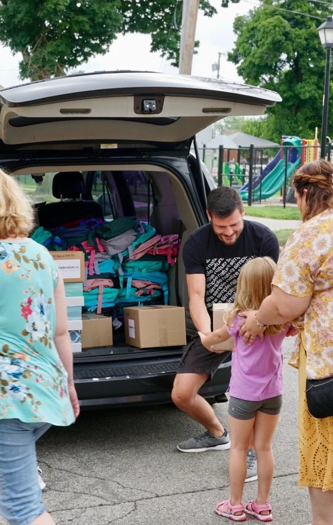 People unloading supplies from a vehicle in a park, including children and adults, with a playground in the background.