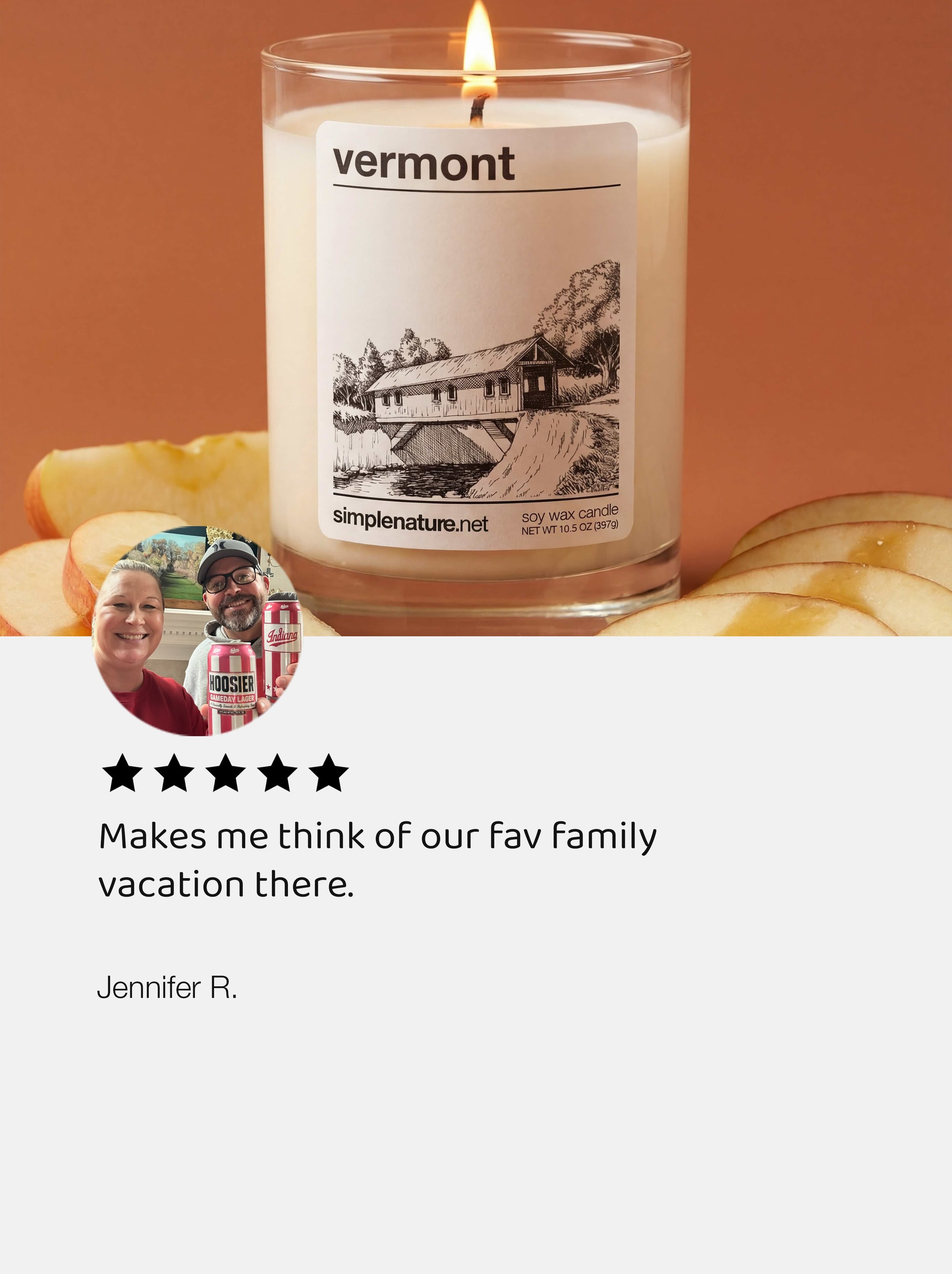 A lit soy wax candle with a black and white illustration of a house on a river, surrounded by trees, labeled 'Vermont' on a warm-colored background. There are sliced apples and a person holding a beer can in the lower left corner.