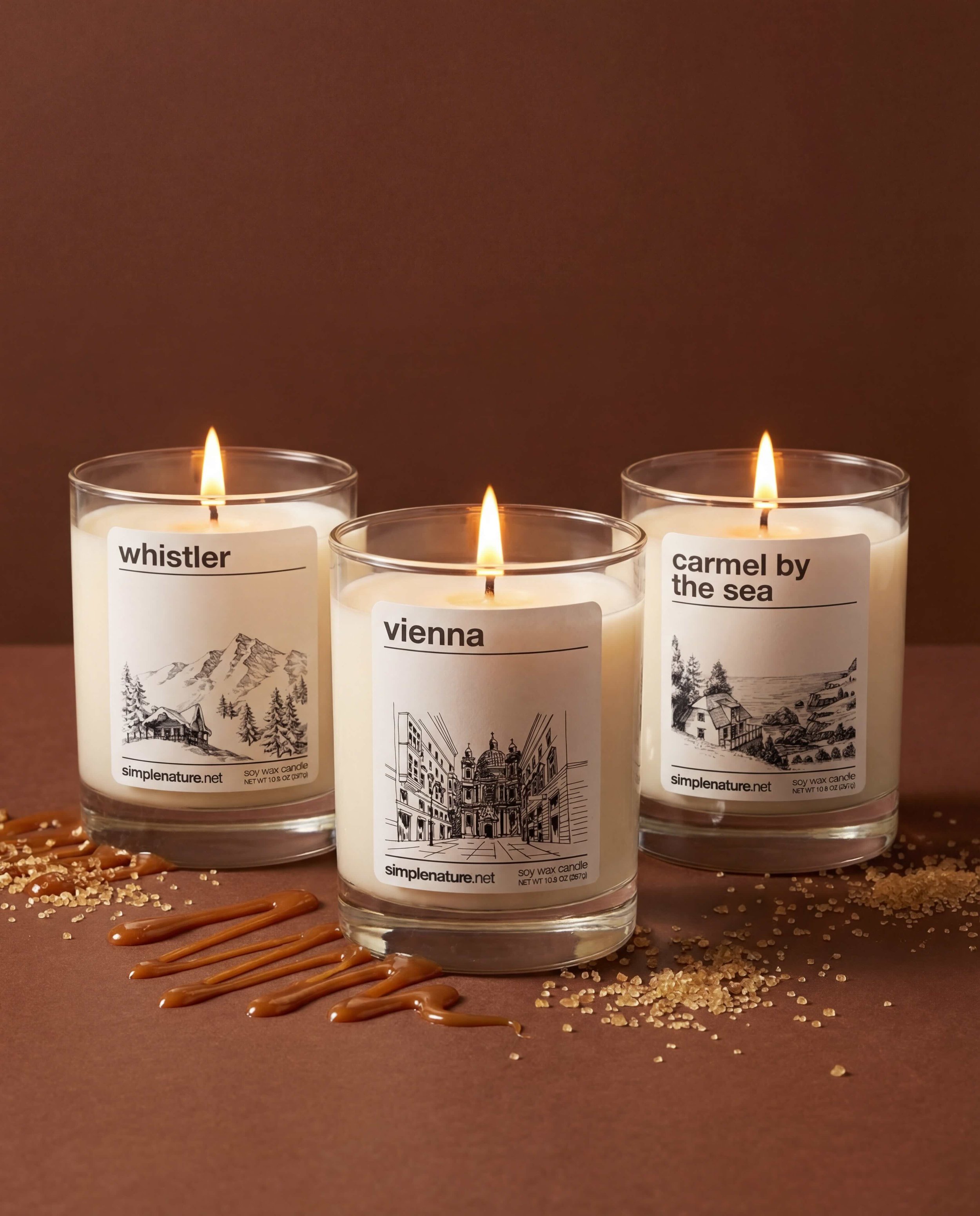 Three lit scented candles in glass containers with nature-themed labels, placed on a wooden surface sprinkled with white powder and sticky orange syrup.