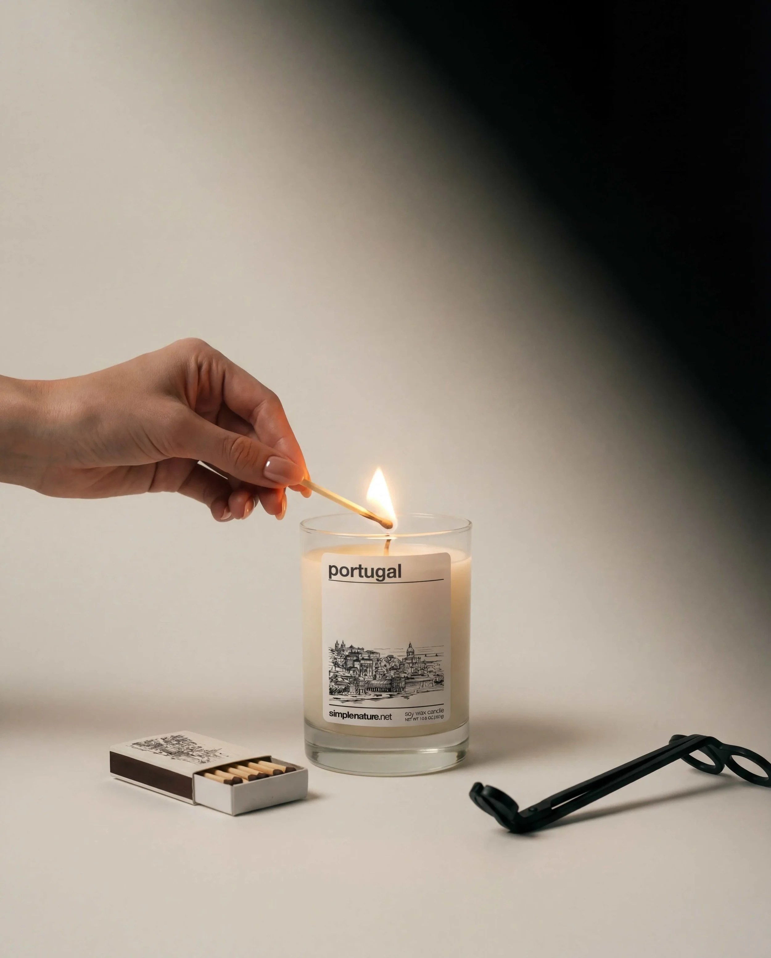 Hand lighting a candle with a matchstick. The candle has a label that reads 'Portugal' and depicts a scenic illustration. Nearby are a box of matches and a candle wick trimmer on a white surface against a gradient background.