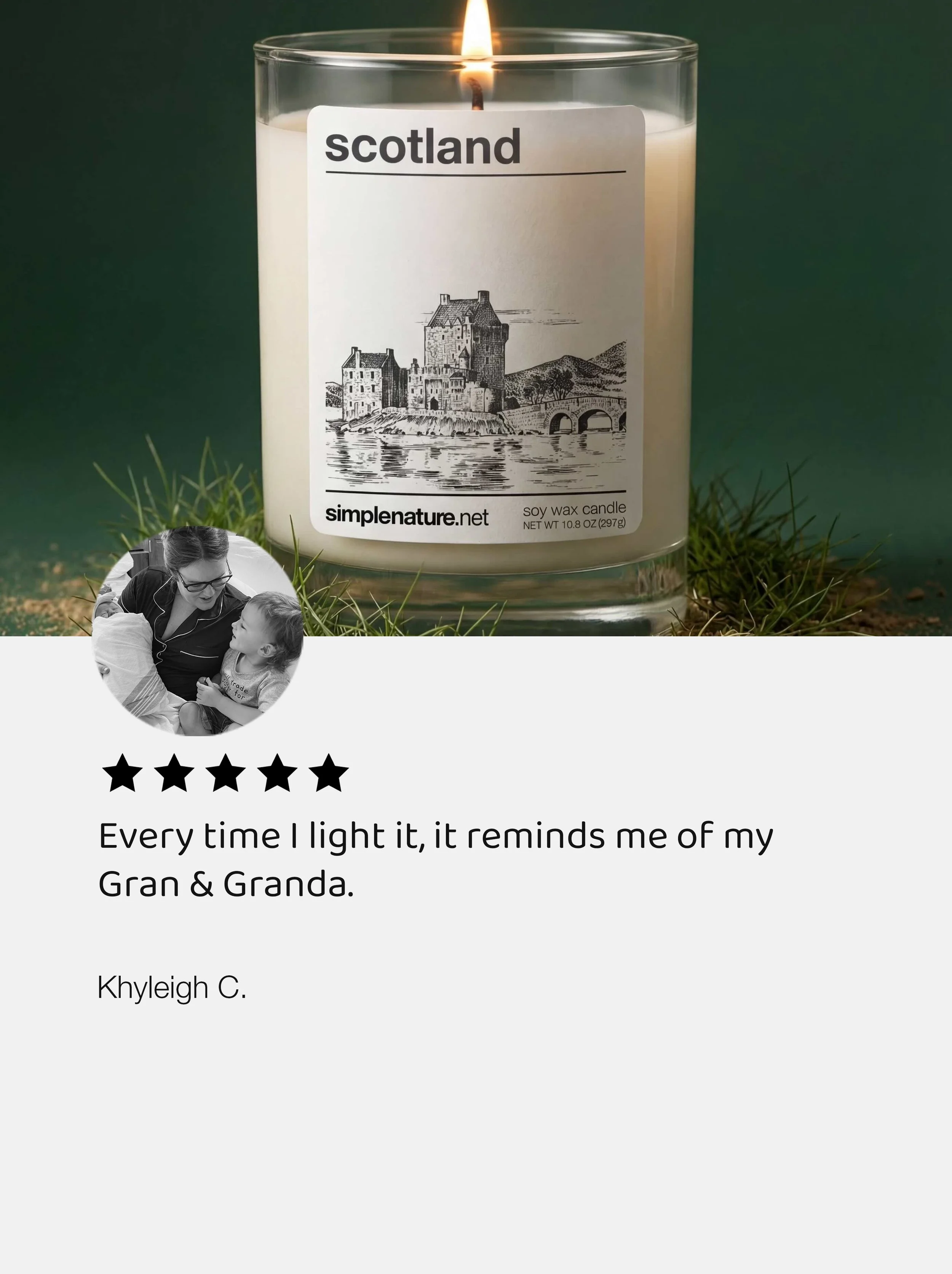 A white candle in a clear glass holder with a label that says "Scotland" and features a drawing of a castle over water. The candle is burning with a small flame. The scene is surrounded by green grass.
