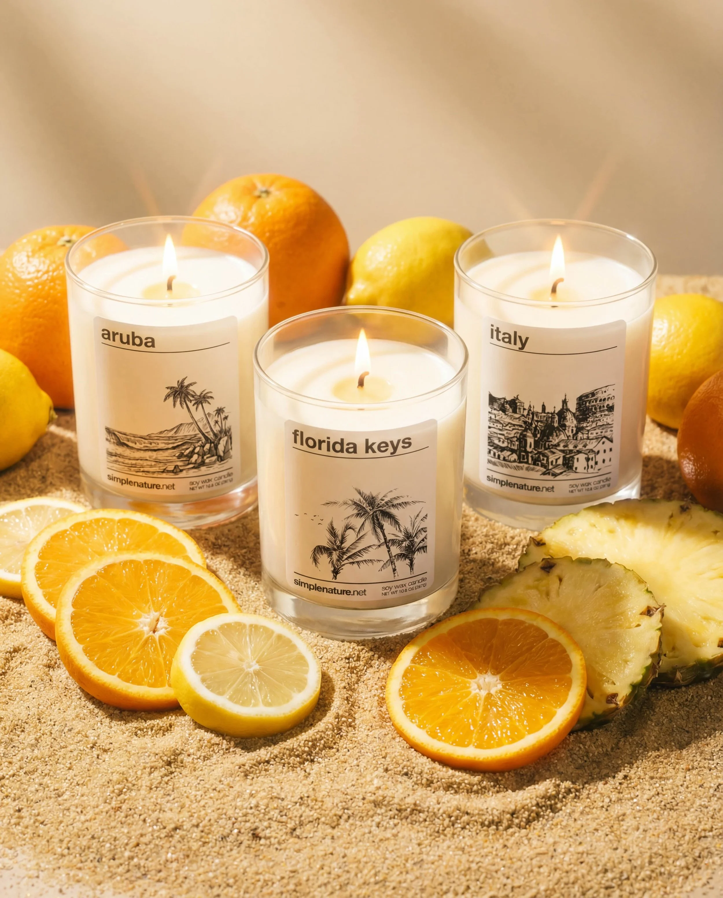 Three lit candles with tropical island labels, surrounded by sliced oranges, lemons, and pineapple on sand.