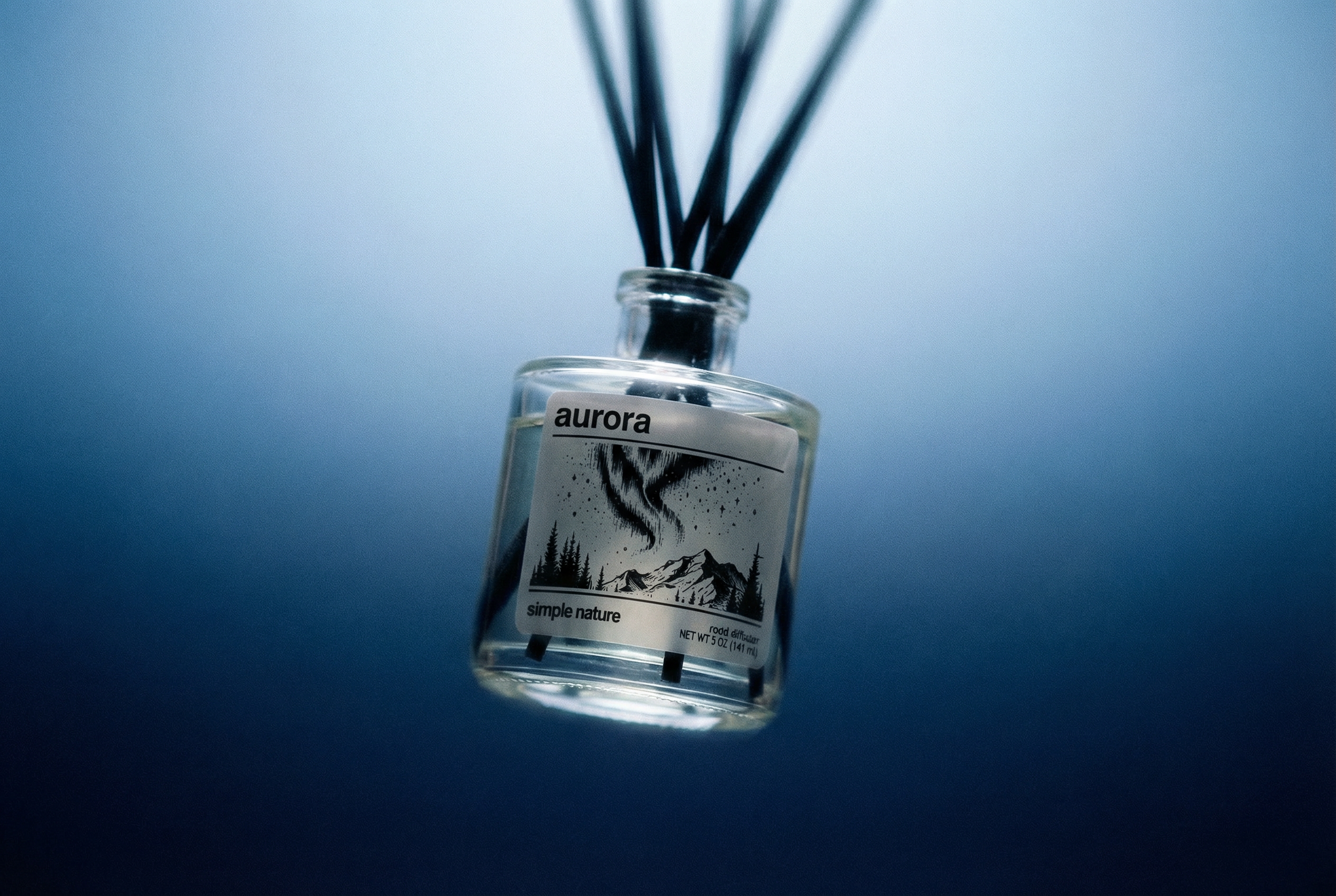A glass diffuser bottle labeled 'aurora' with an illustration of mountains, trees, and a shooting star, containing black reeds, set against a blue background.