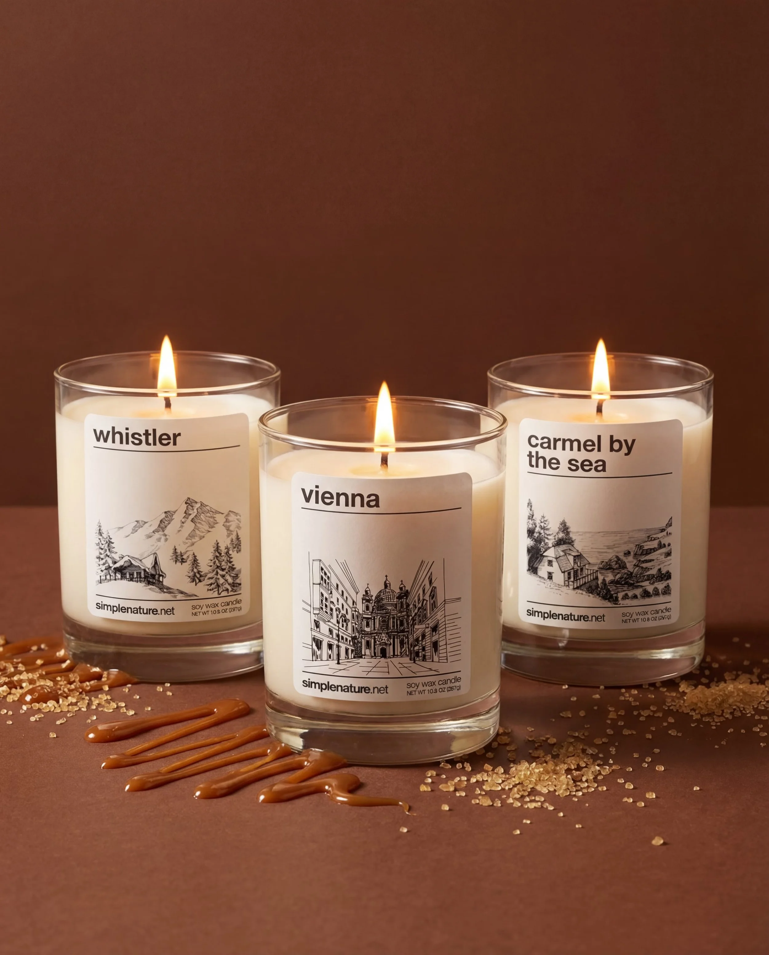 Three lit scented candles in glass containers with nature-themed labels, placed on a wooden surface sprinkled with white powder and sticky orange syrup.
