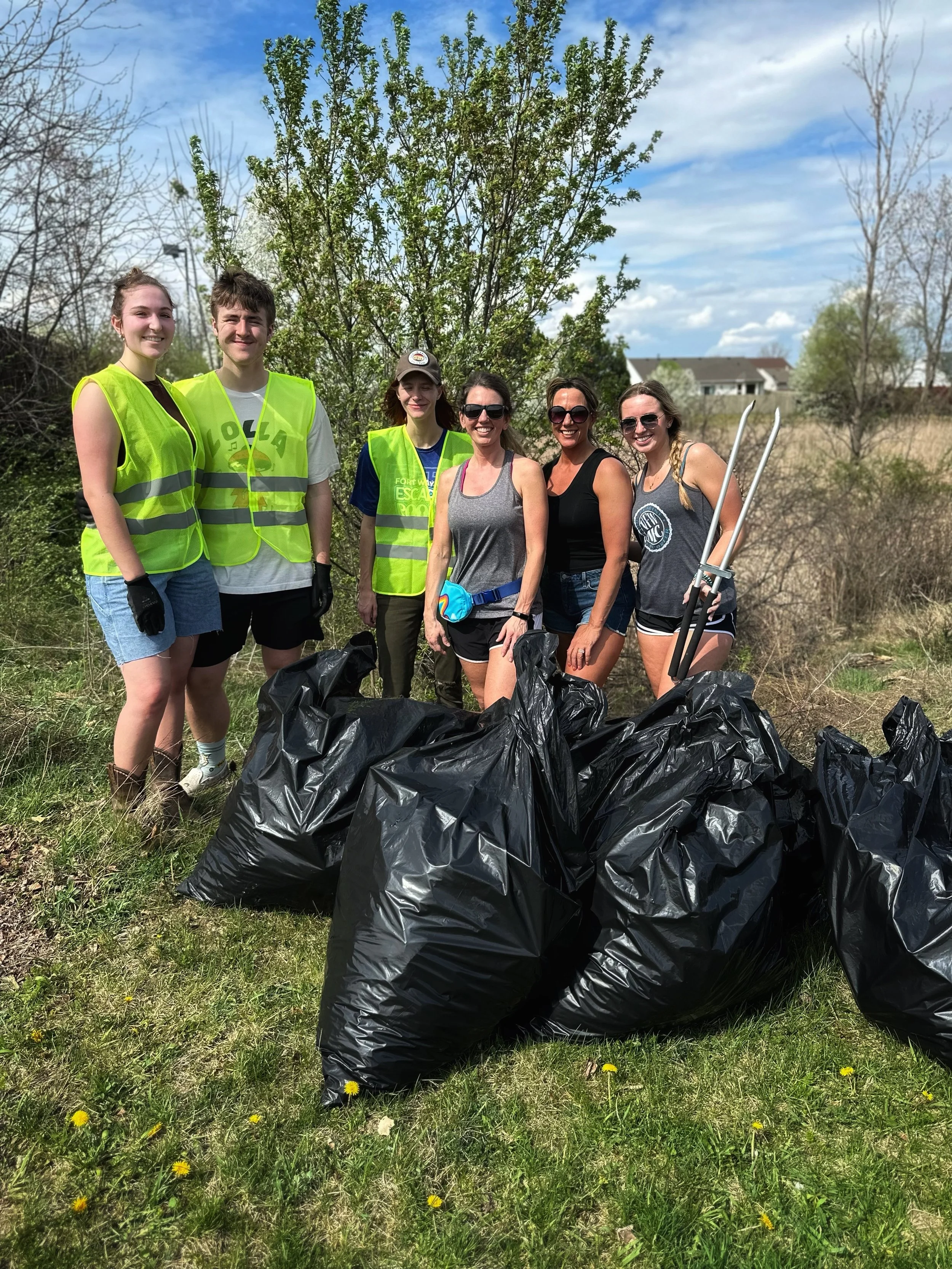 Group of six people, some wearing yellow safety vests, standing outdoors on a grassy area with green trees behind them, after a cleanup event with several black trash bags in front of them.