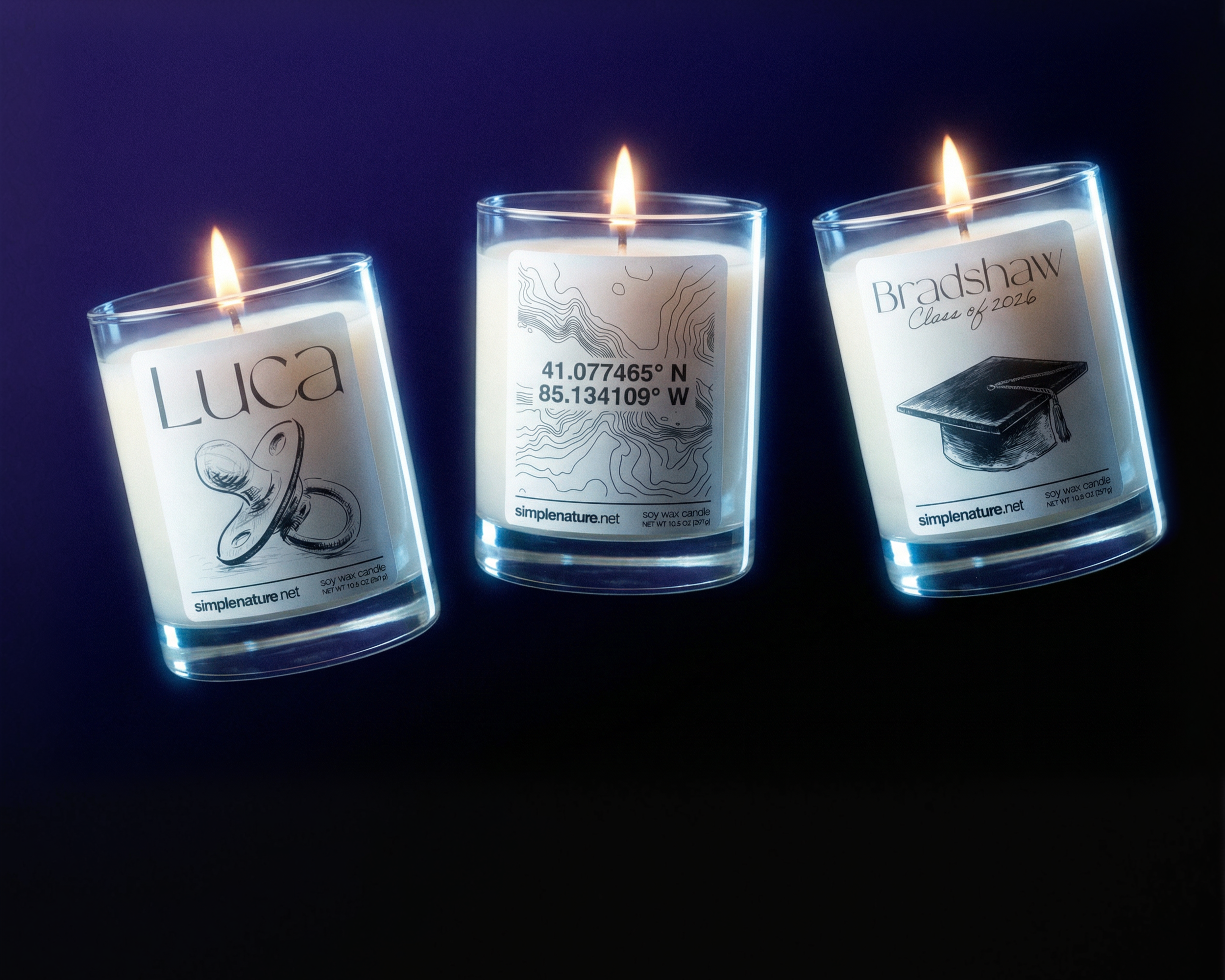 Three lit soy wax candles in glass containers with custom labels, featuring sketches and text, set against a dark background.
