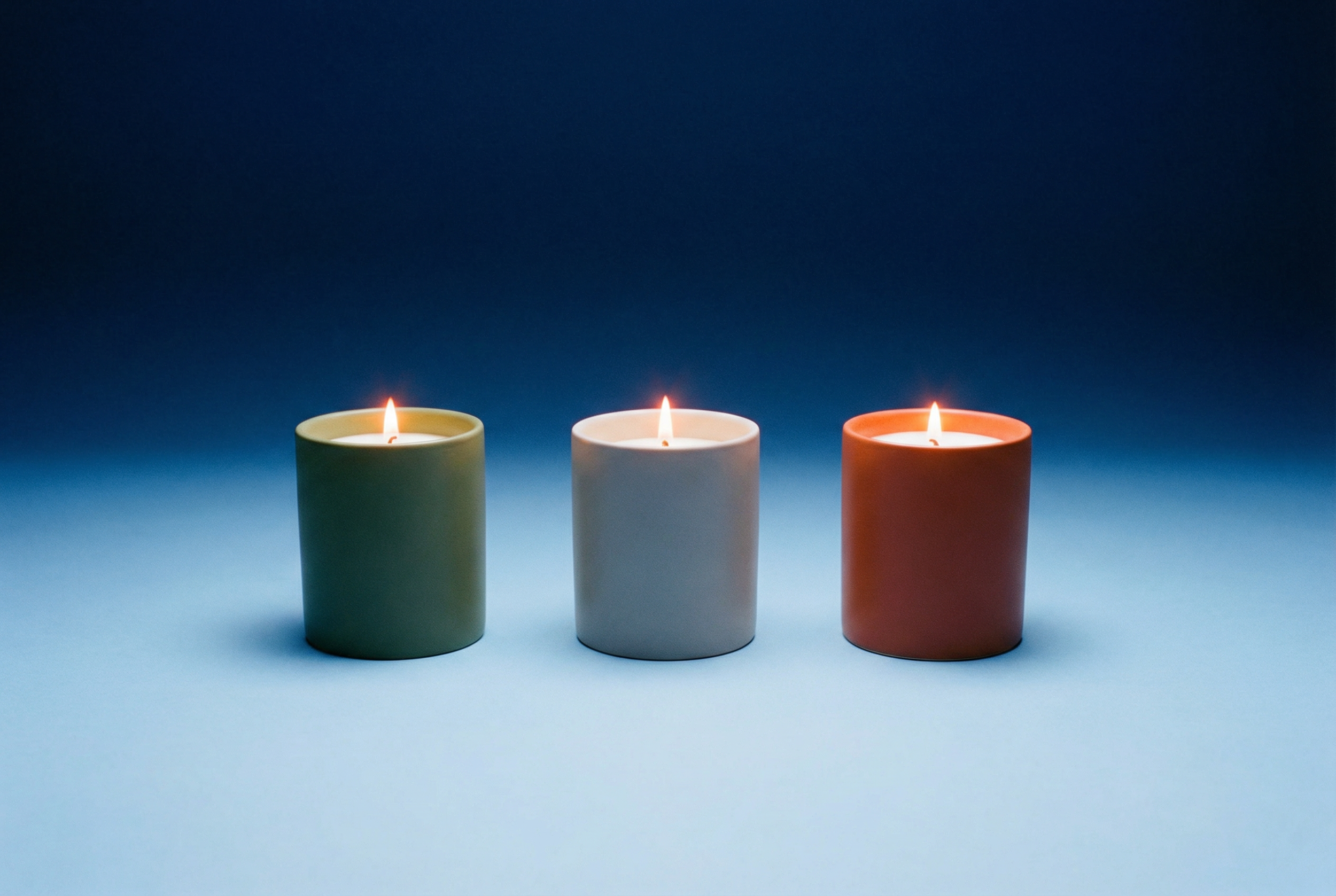 Three lit candles, one green, one white, and one orange, on a surface with a dark blue to light blue gradient background.