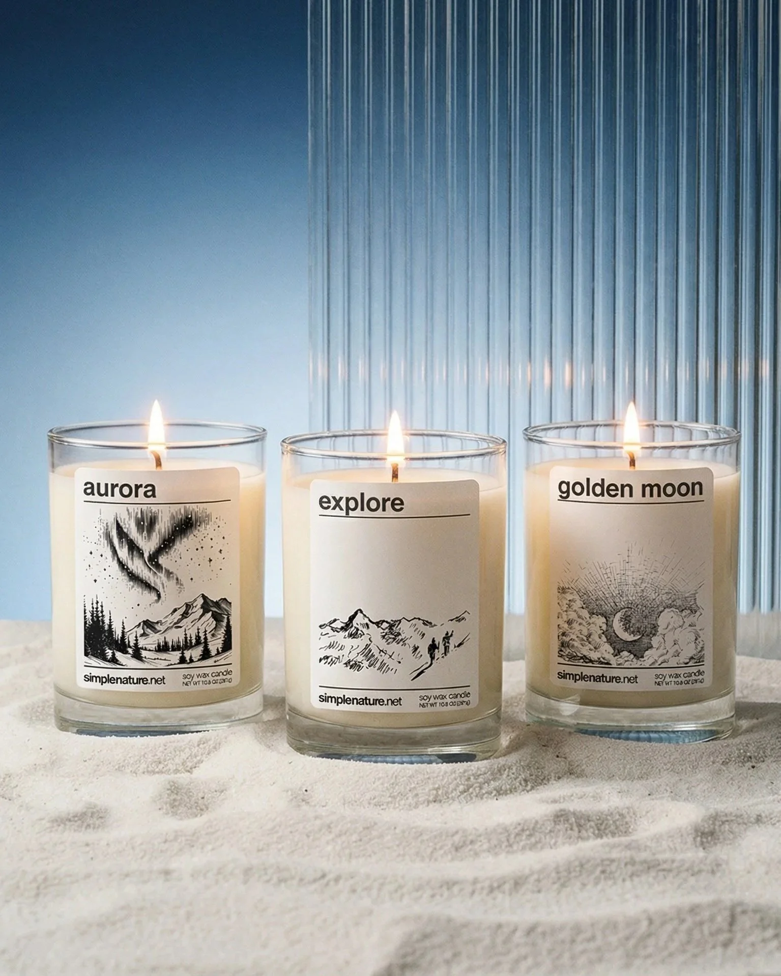 Three white candles with black and white labels reading 'aurora', 'explore', and 'golden moon', lit and placed on a sandy surface with a blue background and vertical textured panel.