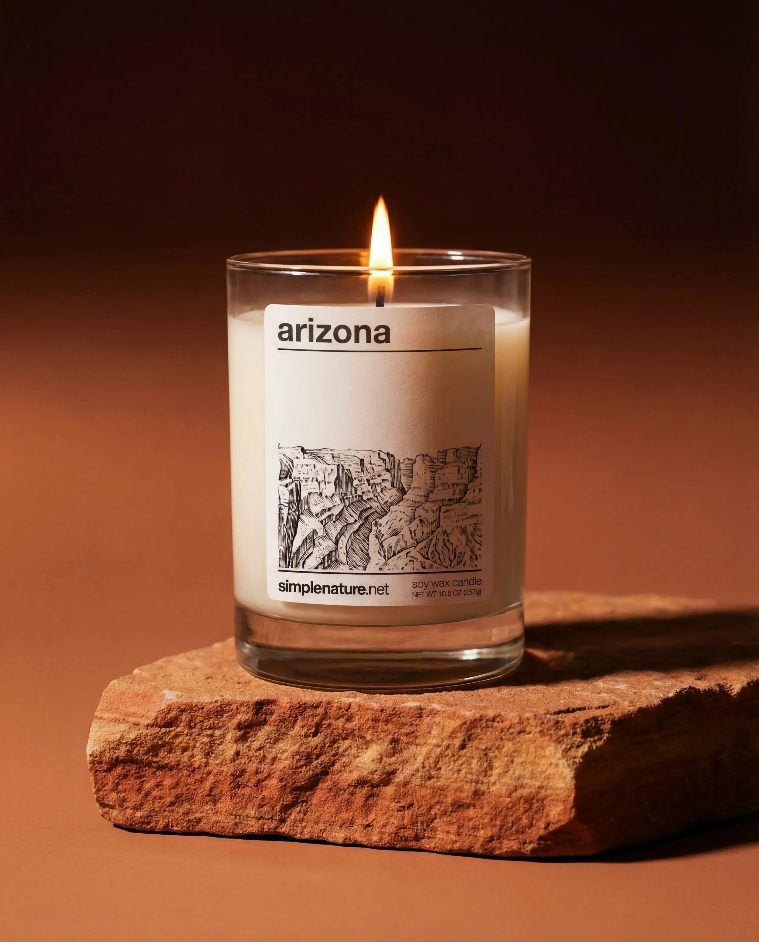 A lit candle in a glass jar labeled Arizona, sitting on a piece of rough, reddish stone against a dark, warm background.