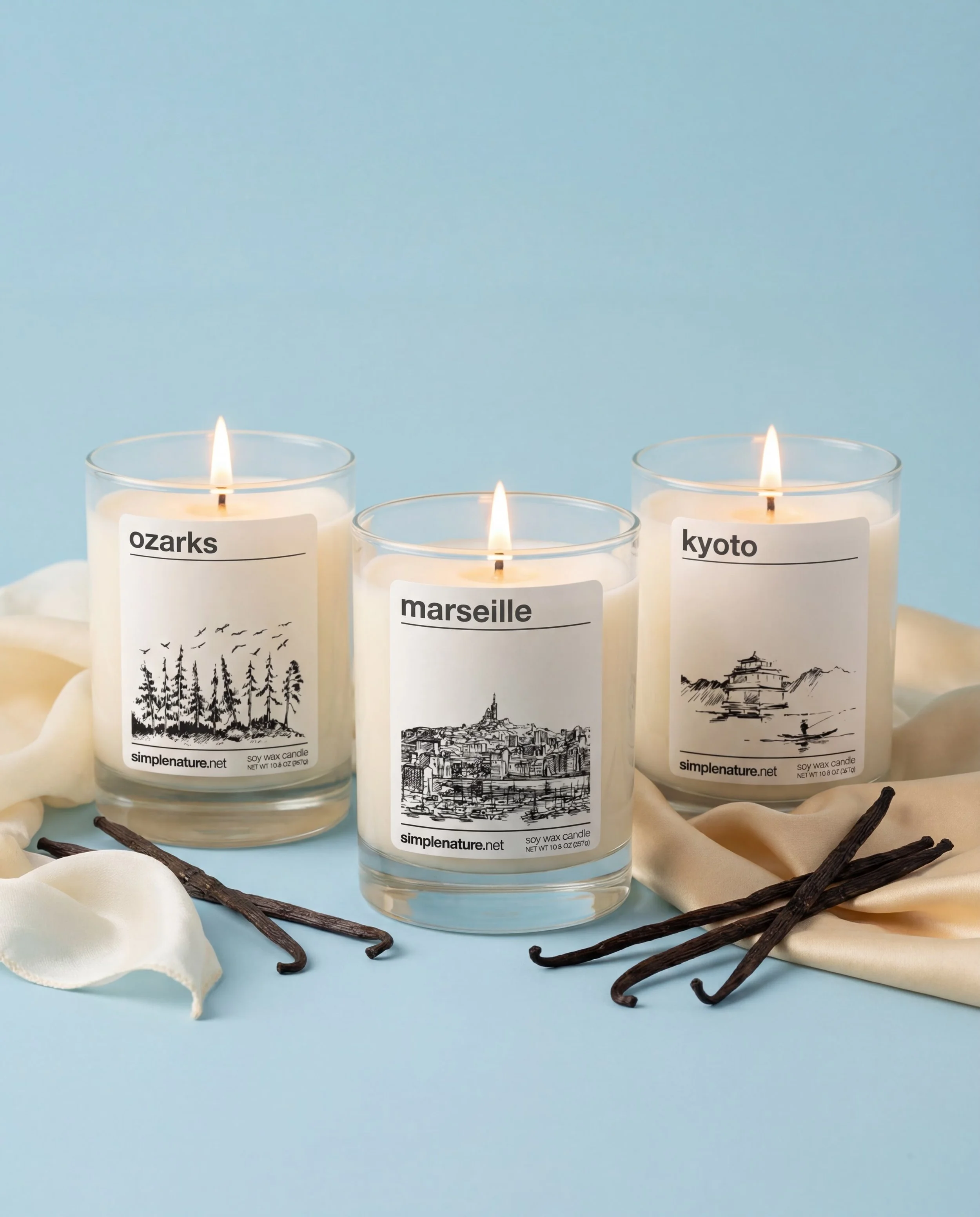 Three lit candles in glass containers with nature-themed labels reading 'aurora,' 'explore,' and 'golden moon'; they are on a textured white surface with a metallic blue corrugated background.