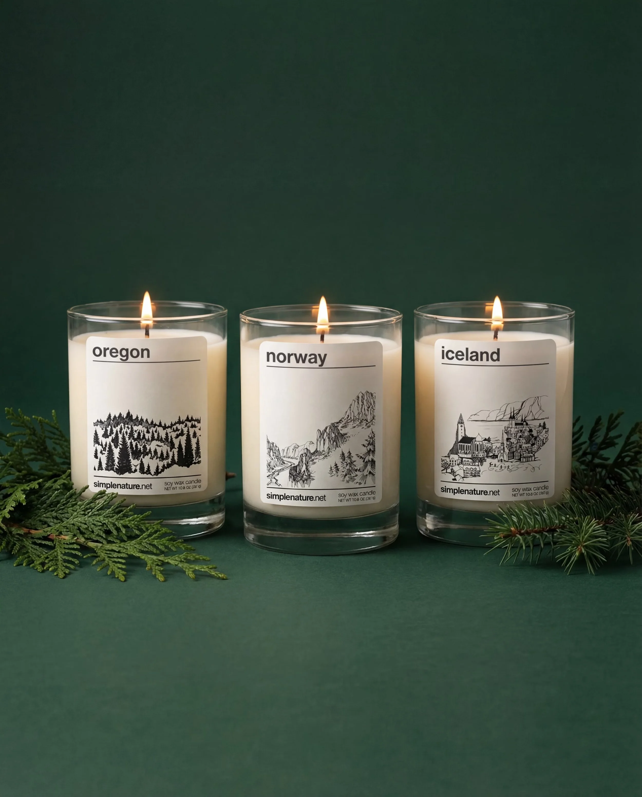 Two lit candles with labels 'Norway' and 'Oregon' on a wooden surface surrounded by pine branches and logs, with a dark background.