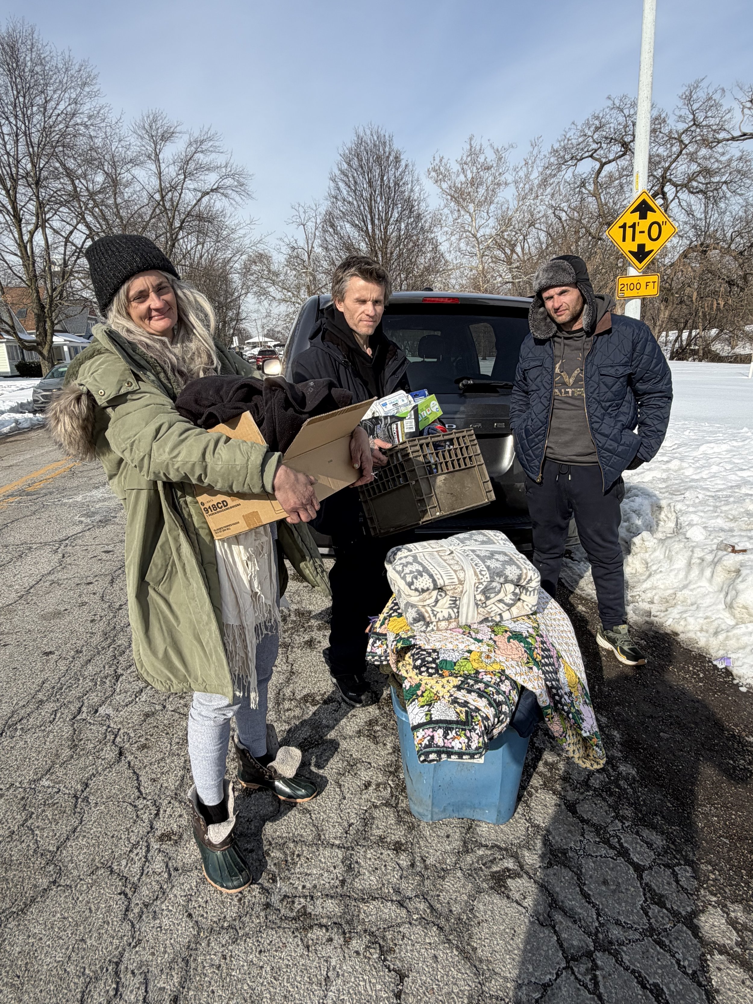 Three people standing outdoors on a snowy day near a black vehicle, with donations including boxes, crates, and blankets, likely for aid or charity.